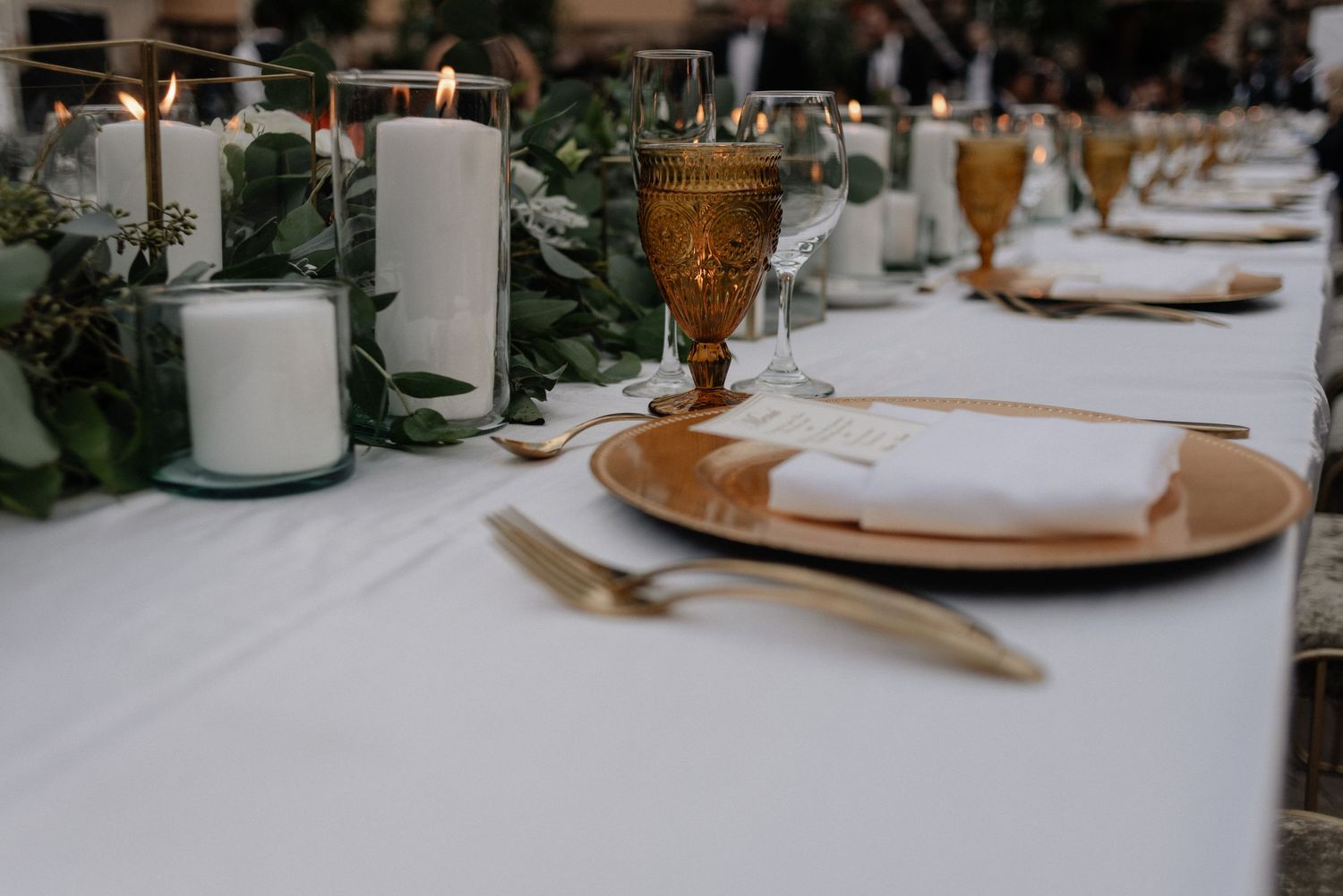 Elegant table setting with amber glassware, white candles, greenery and gold charger plates on white tablecloth.