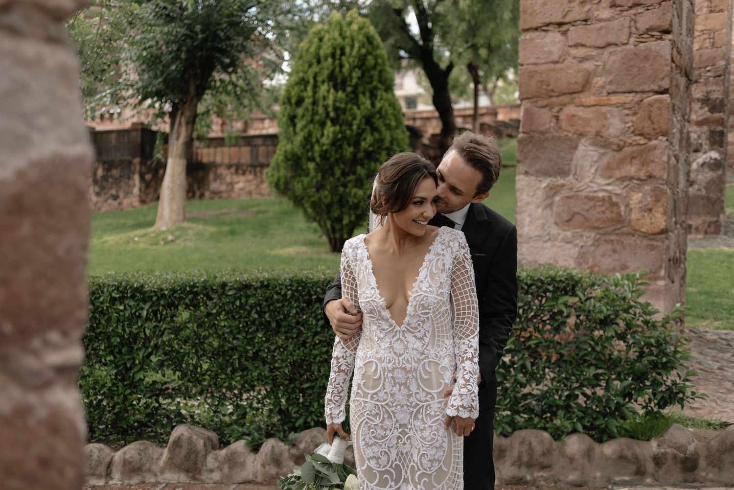 A couple shares an intimate moment in a garden setting against stone walls and green hedges, with the person wearing a beaded gown.