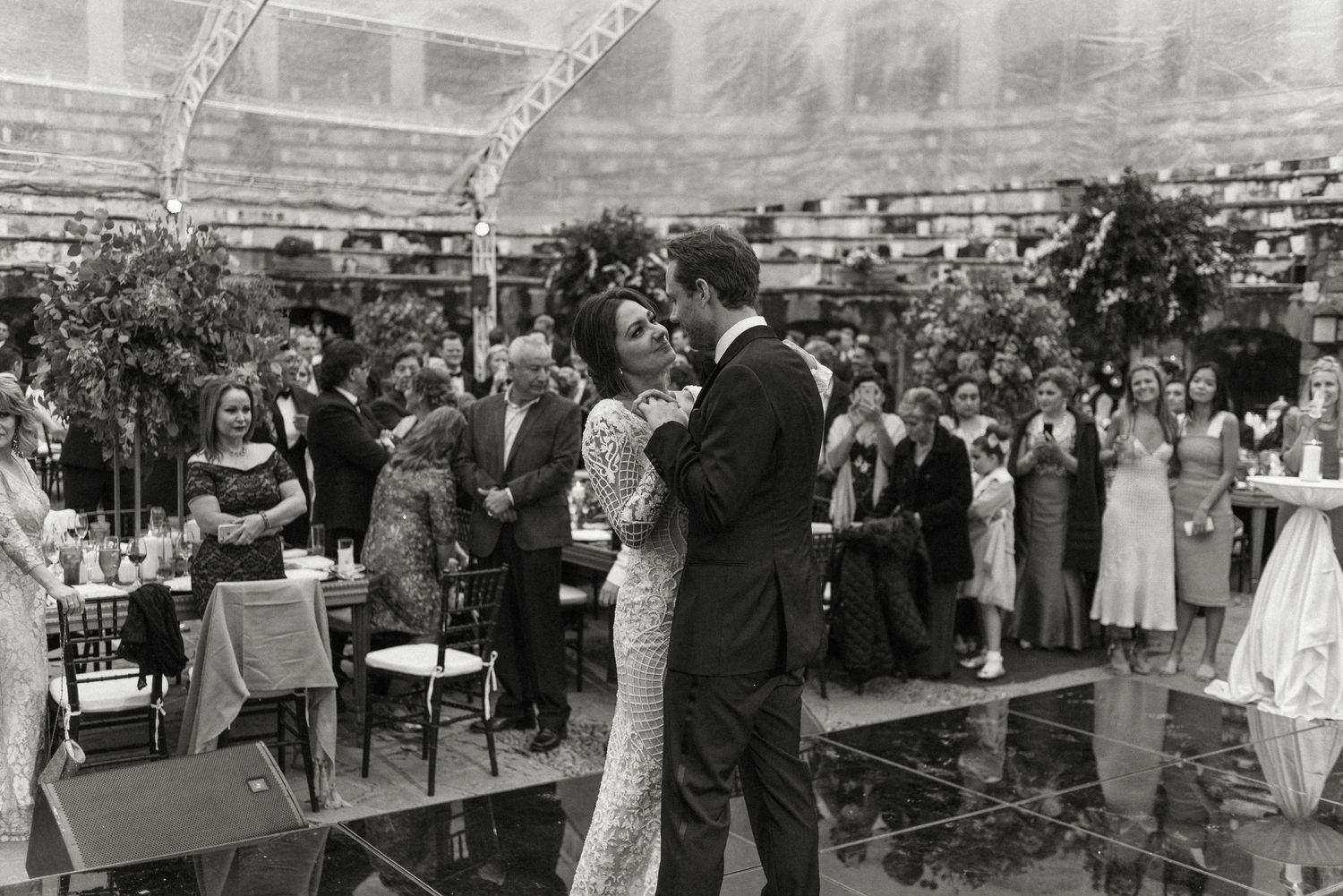 A couple shares their first dance at a wedding reception under a clear tent with guests looking on in black and white photography.