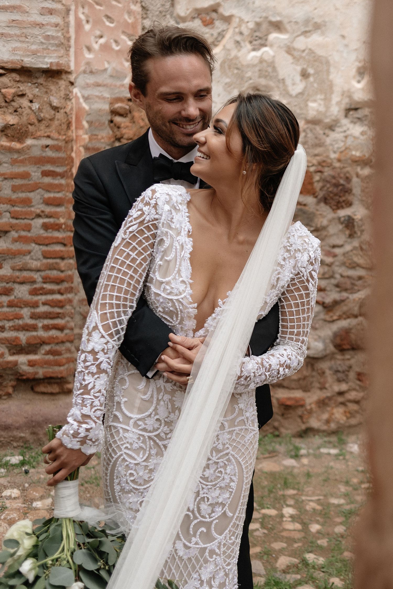 A newlywed couple embraces against a rustic brick wall, with the bride wearing an intricate lace gown and flowing veil.