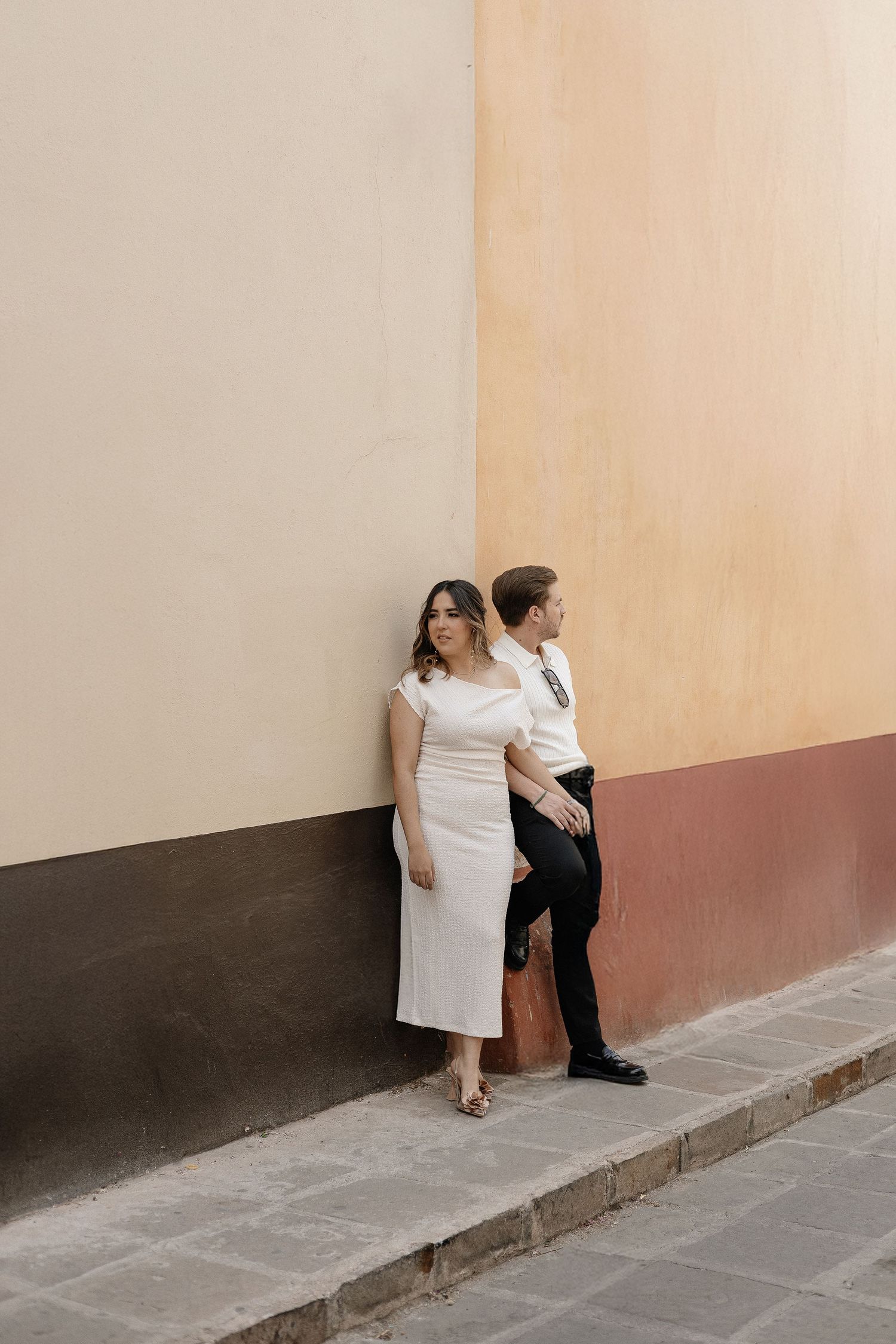 Two people sit close together on a sidewalk against a beige and brown wall in a minimalist urban setting.