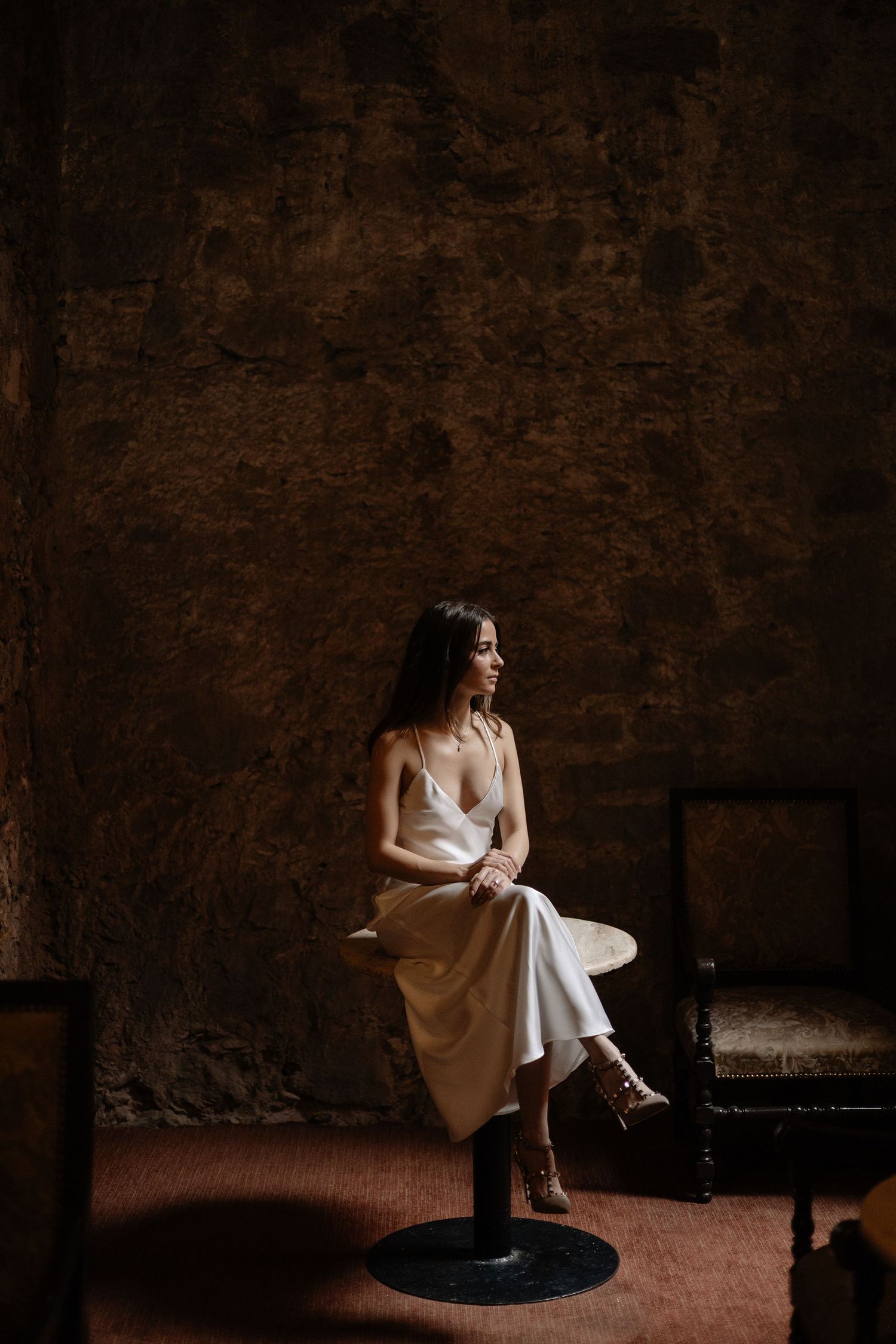 Artistic moody portrait of a bride sitting alone in a dimly lit rustic stone room.