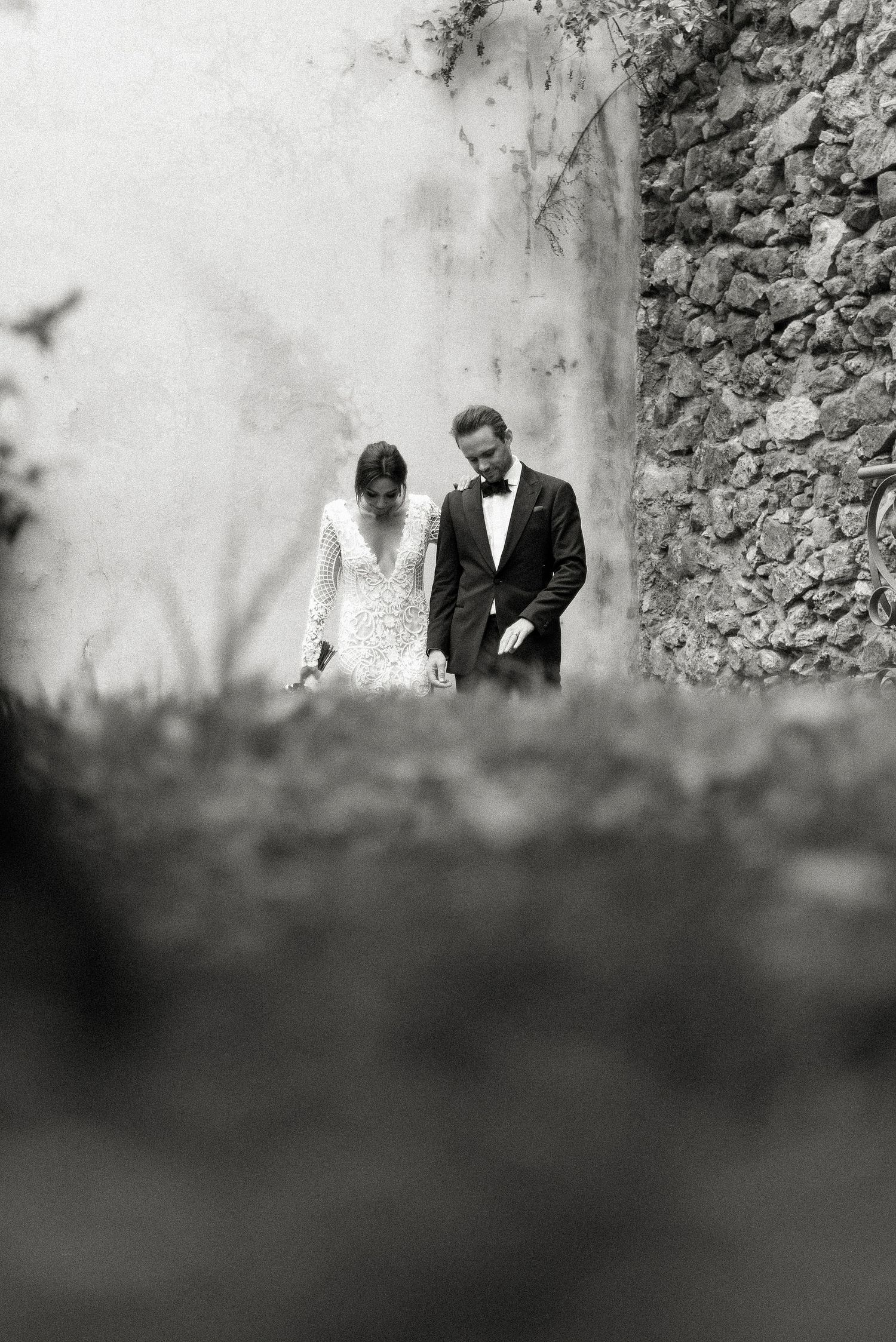 A romantic black and white wedding photograph showing a couple in formal attire standing against a stone wall.