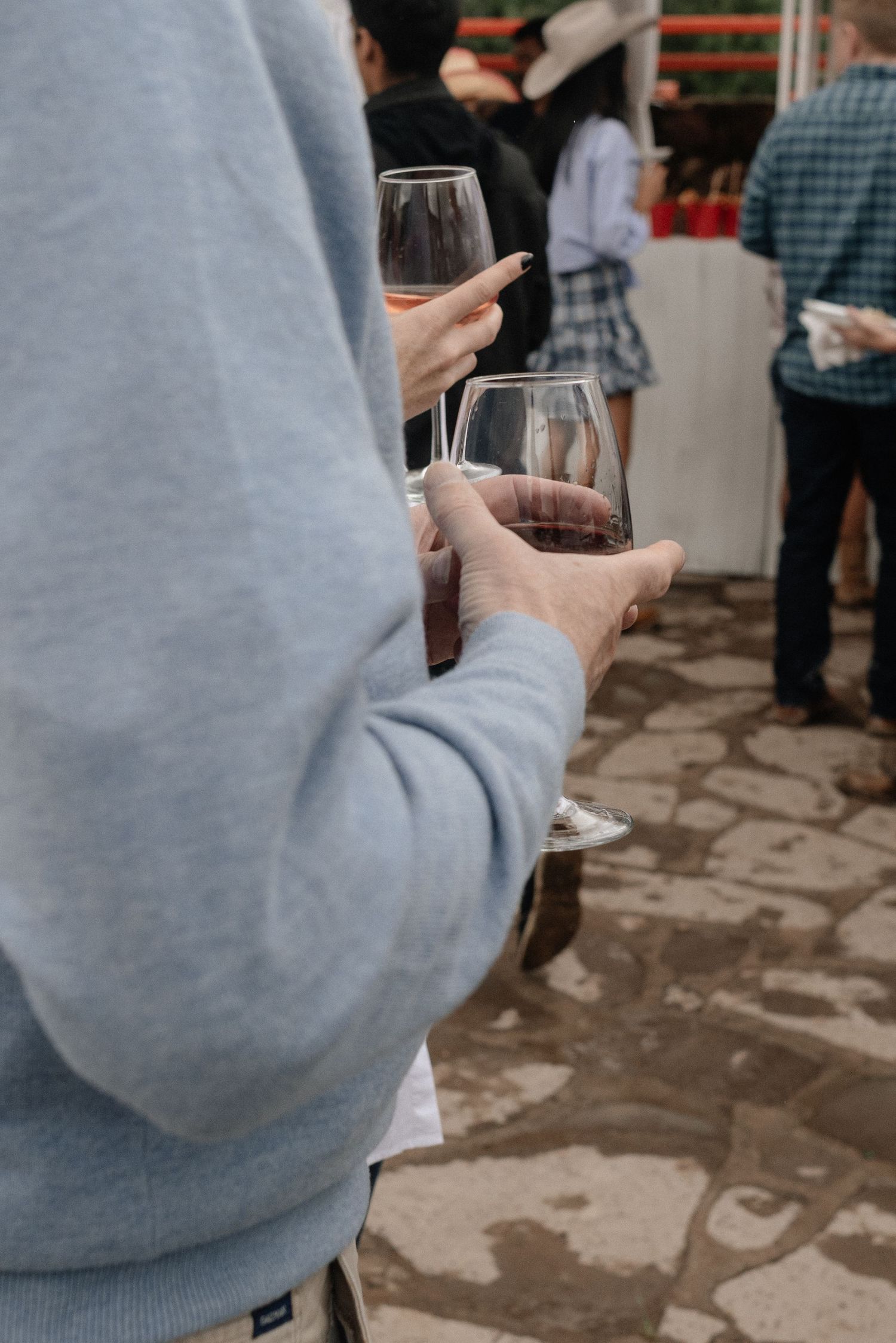 Someone in a gray sweater holds a glass of red wine at a social gathering with people visible in the background.