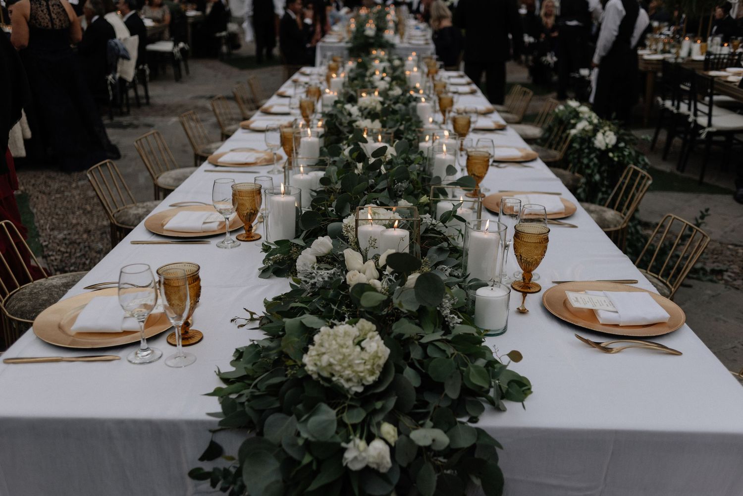 Long wedding reception table with elegant white floral centerpiece, amber goblets and eucalyptus garland runner.