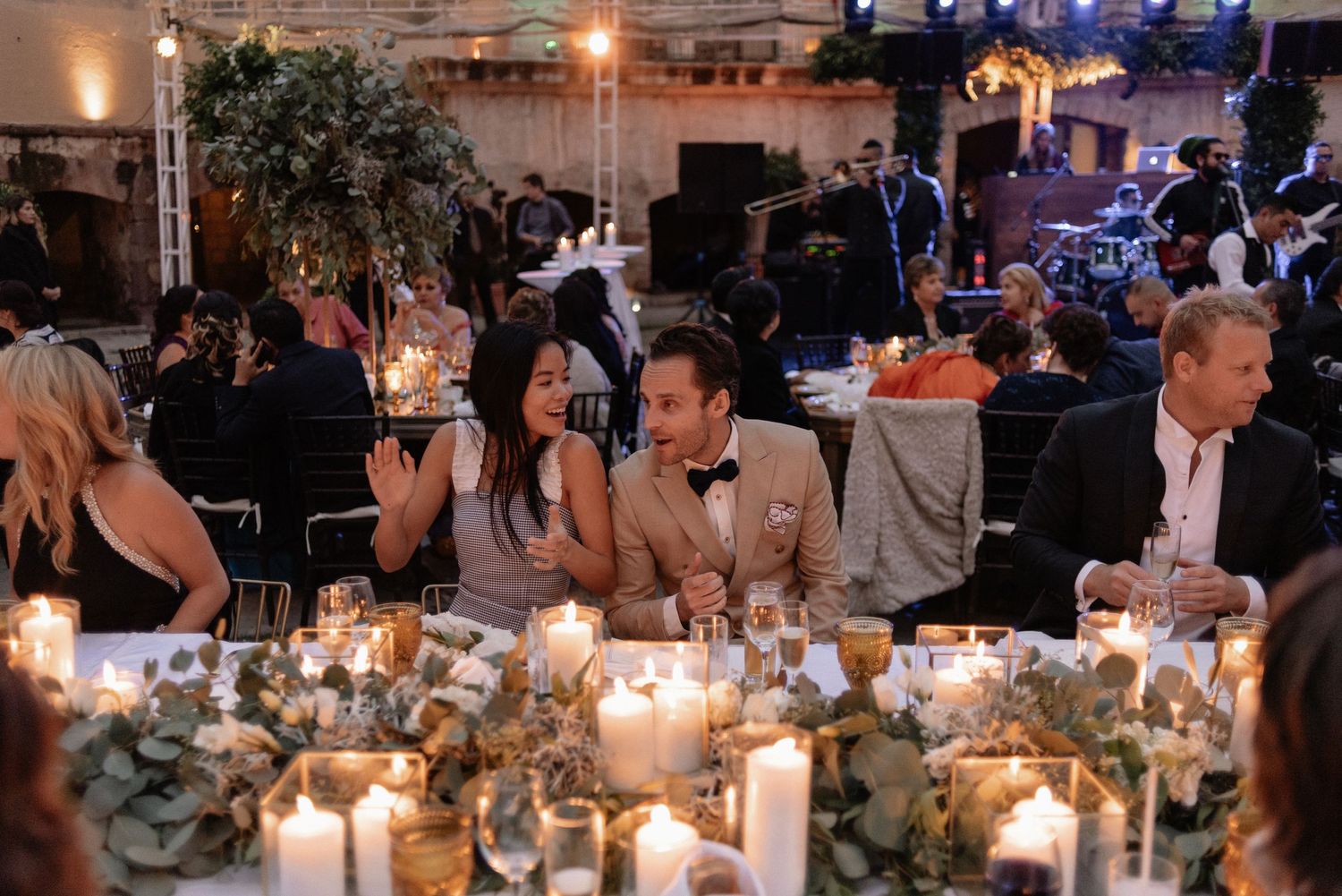 Wedding reception guests sit at a candlelit table decorated with lush greenery and multiple glowing centerpieces.