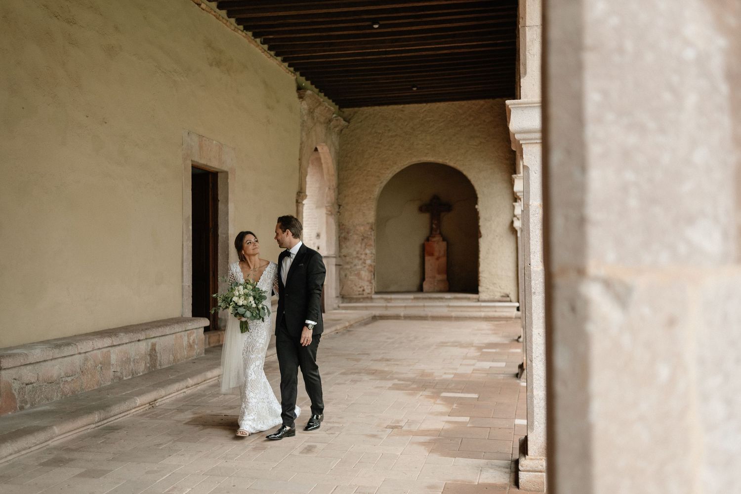 A couple in wedding attire stands in a historic arched hallway with stone columns and white stucco walls.