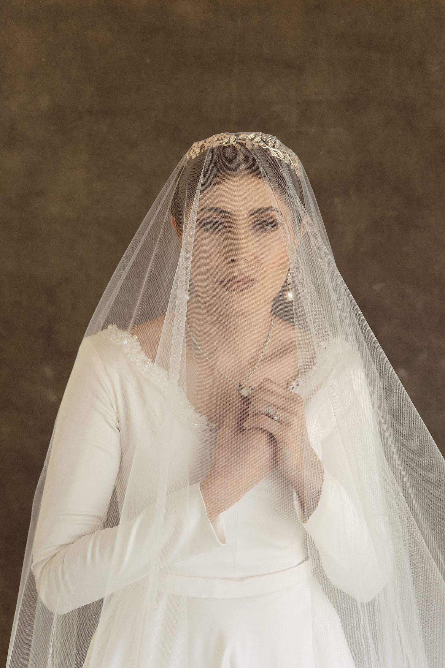 A bride in a classic long-sleeved white wedding dress and veil poses against a dark brown backdrop in vintage style.