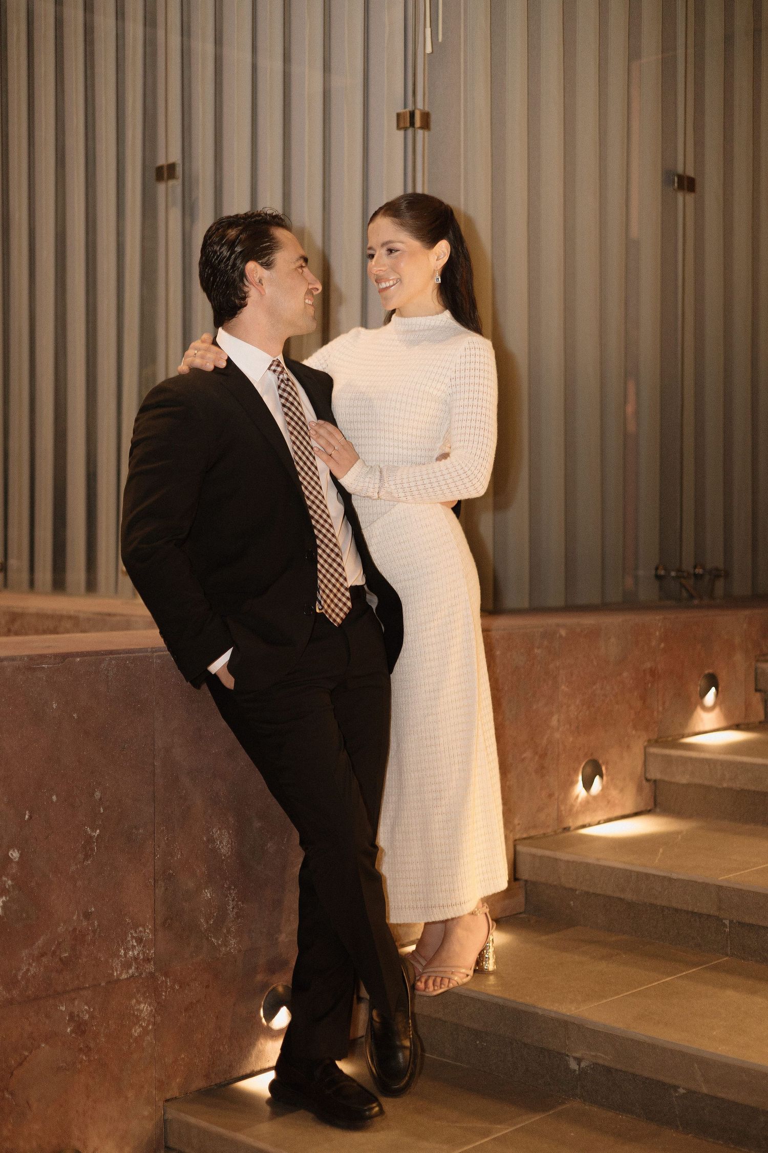 A couple in formal attire share an intimate moment on marble steps, with the woman in white and man in dark suit.