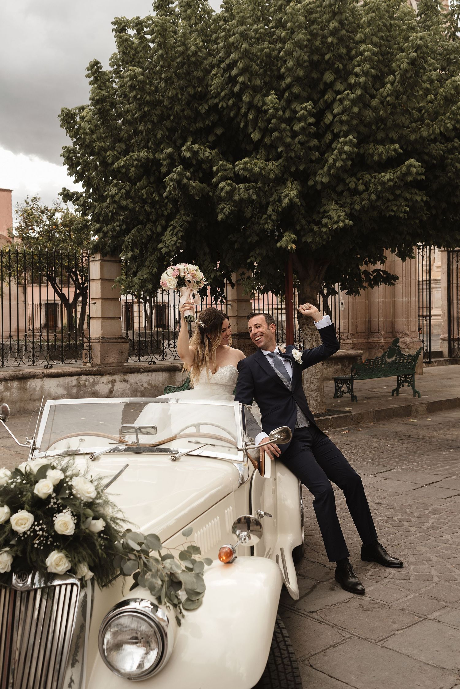 Classic white vintage car decorated with flowers serves as elegant wedding transportation on a cobblestone street.