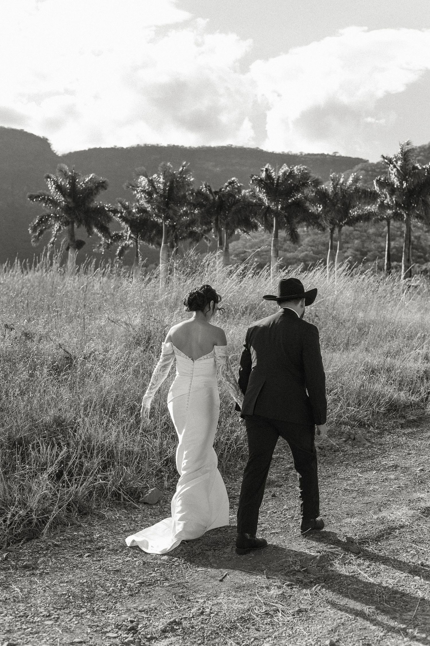 A couple in wedding attire sits together on a grassy hillside overlooking a scenic tropical landscape with palm trees.