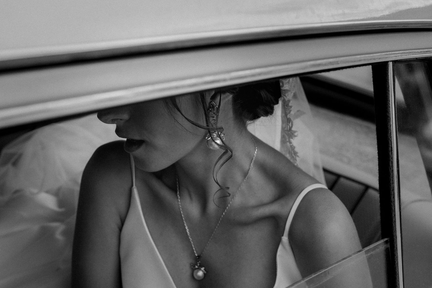 An artistic black and white portrait of a bride wearing jewelry while seated in a car.