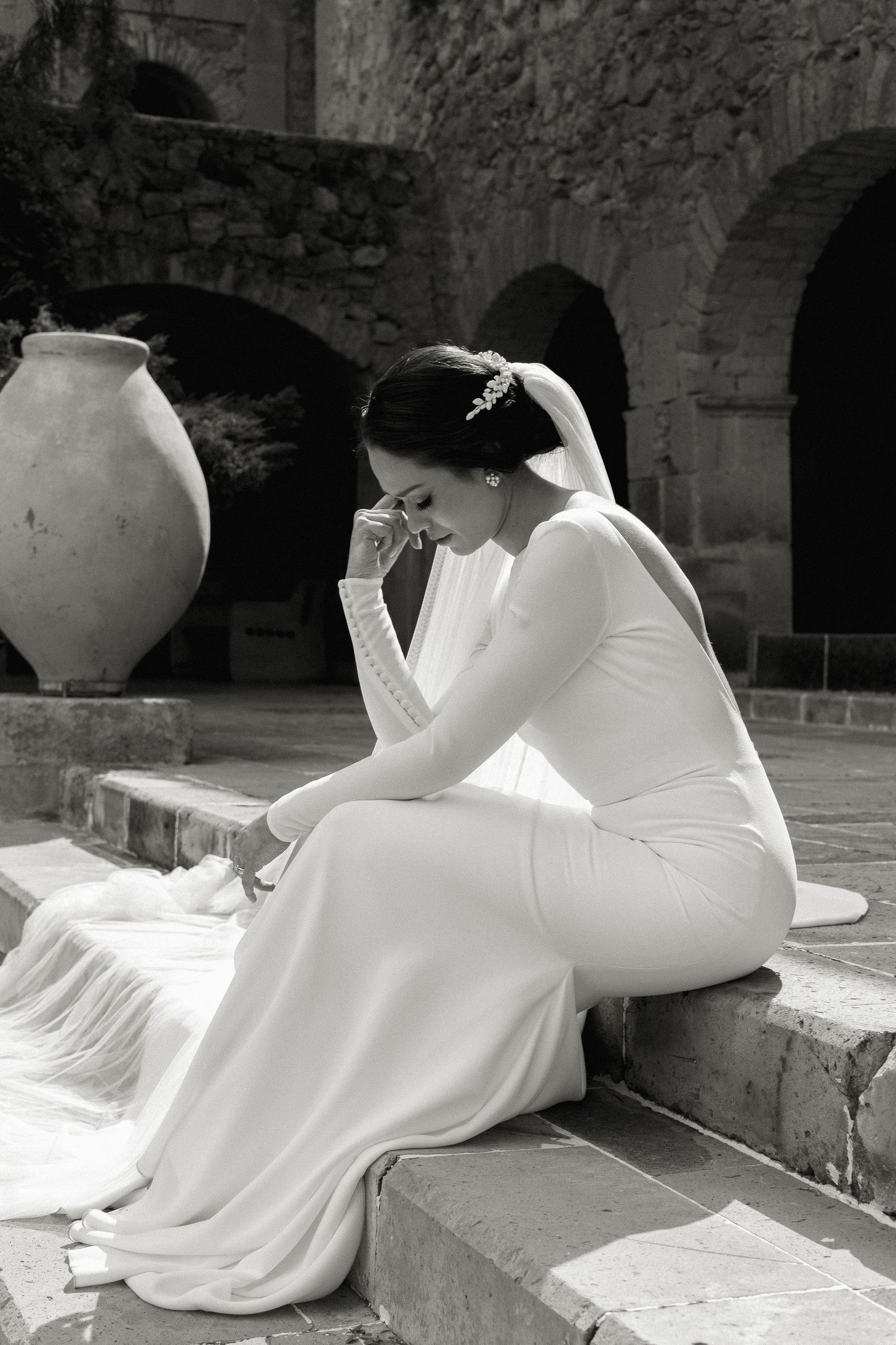 Black and white photo of bride in elegant dress seated by large clay vessel.