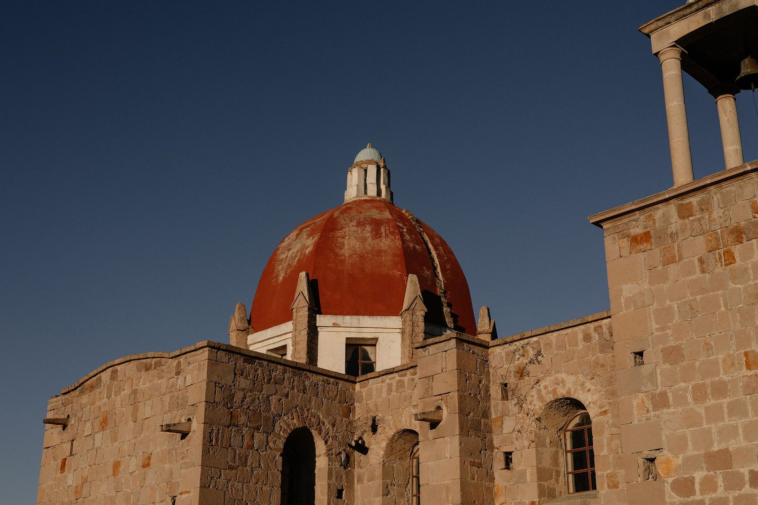 Historic stone church buildings with red domed roofs stand against a clear blue sky in a Mediterranean setting.