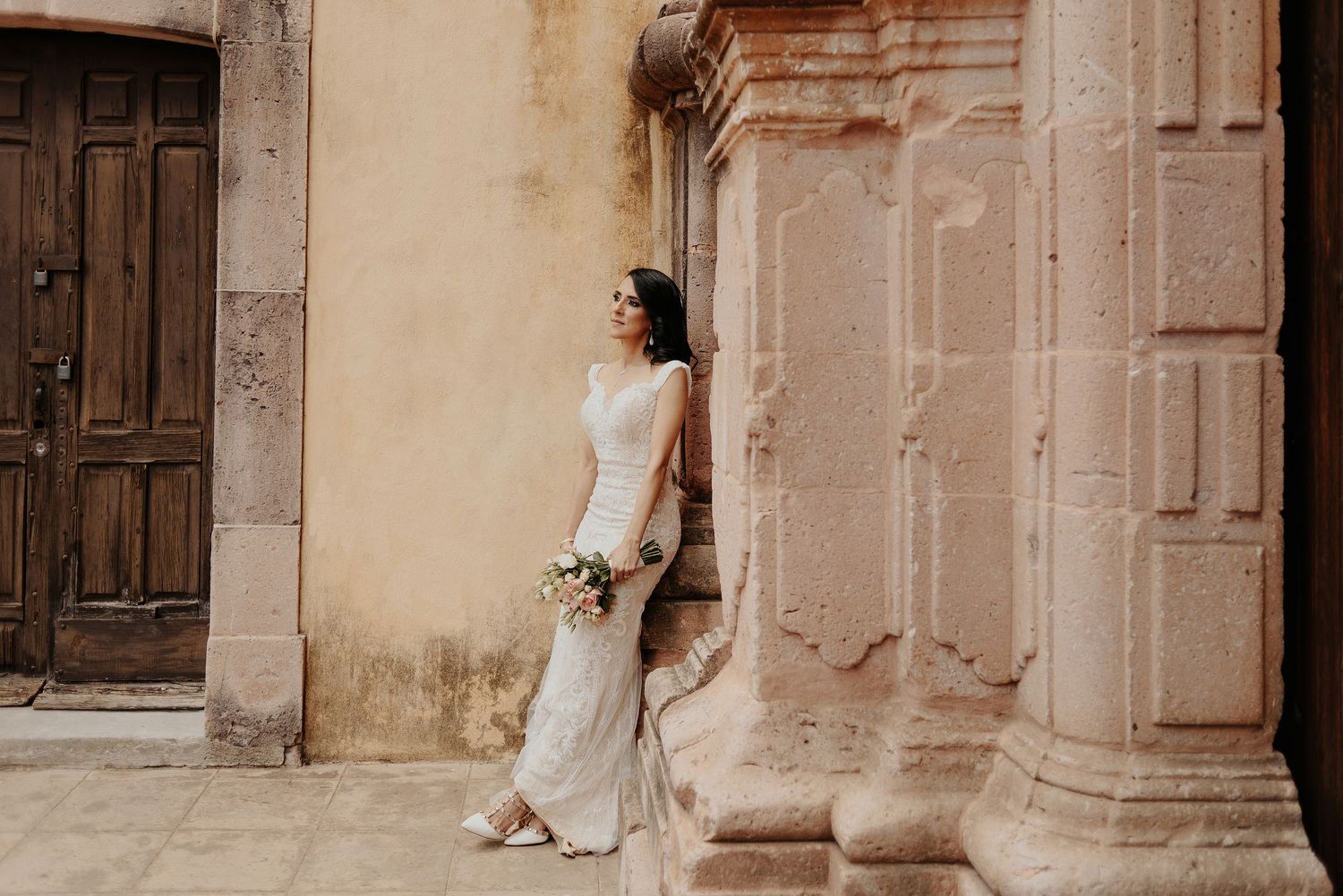 Bride in elegant white dress poses against rustic stone wall with architectural details.
