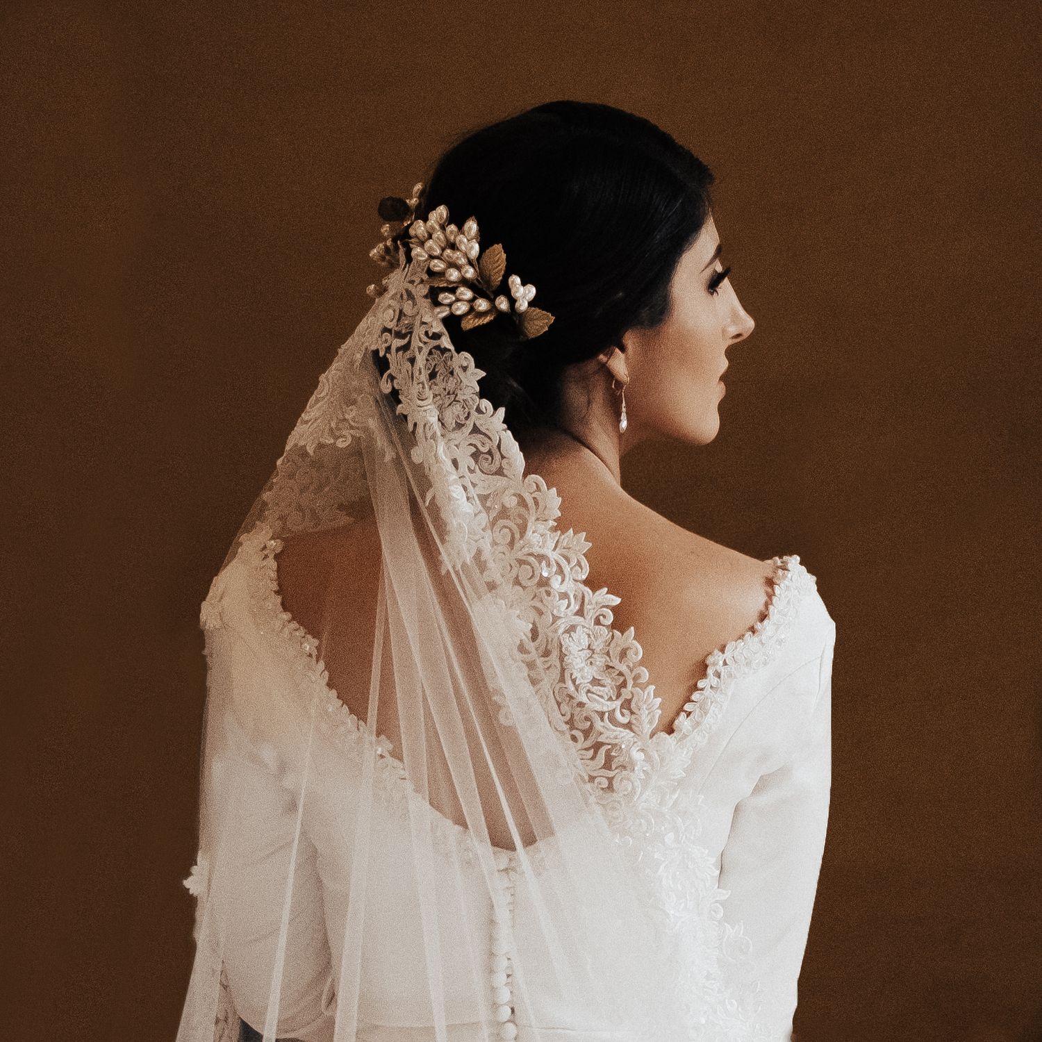 Artistic profile view of a bride wearing a delicate lace veil against a rich brown background.