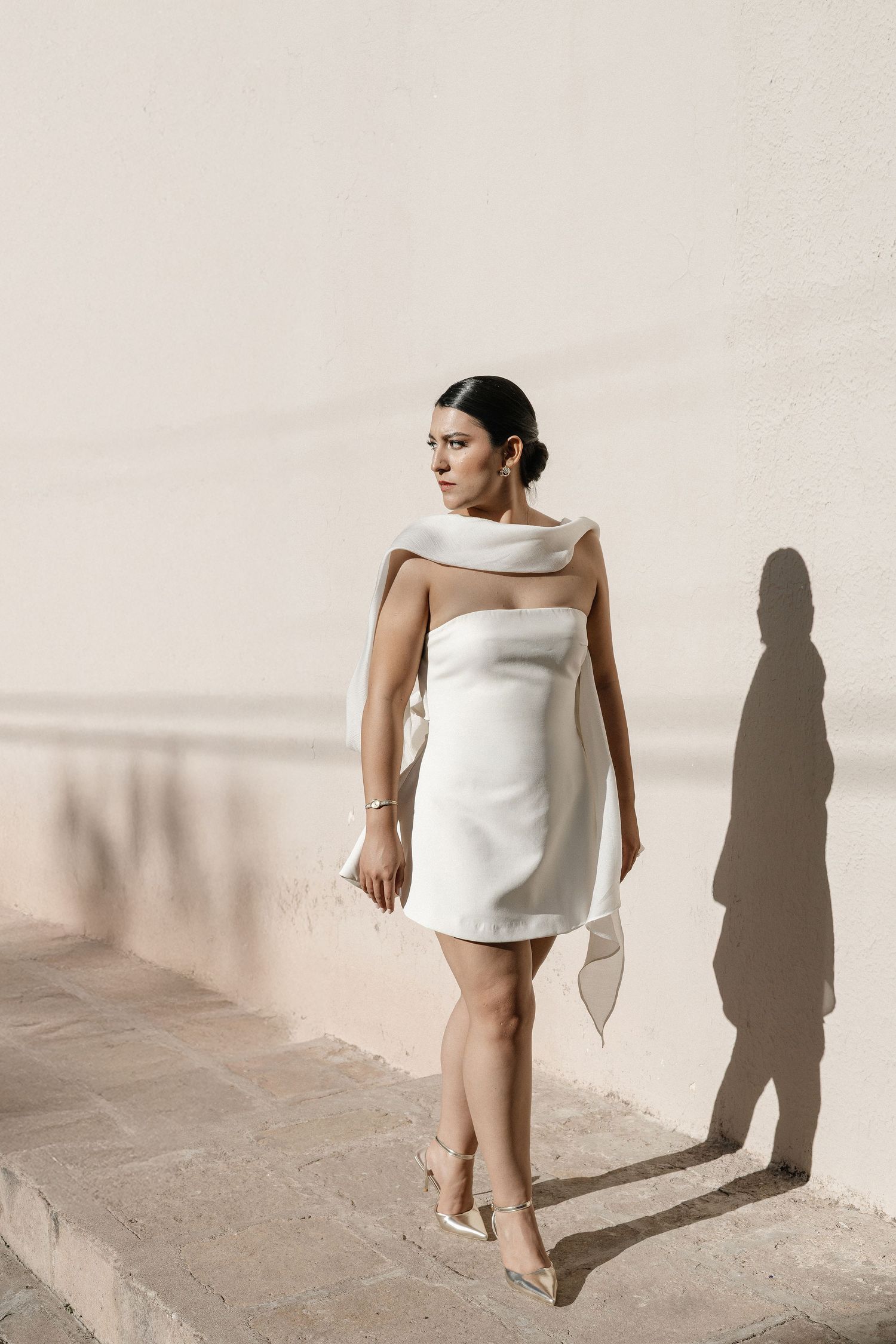 A person in a white mini dress poses against a light wall while casting dramatic shadows in a minimalist photoshoot series.
