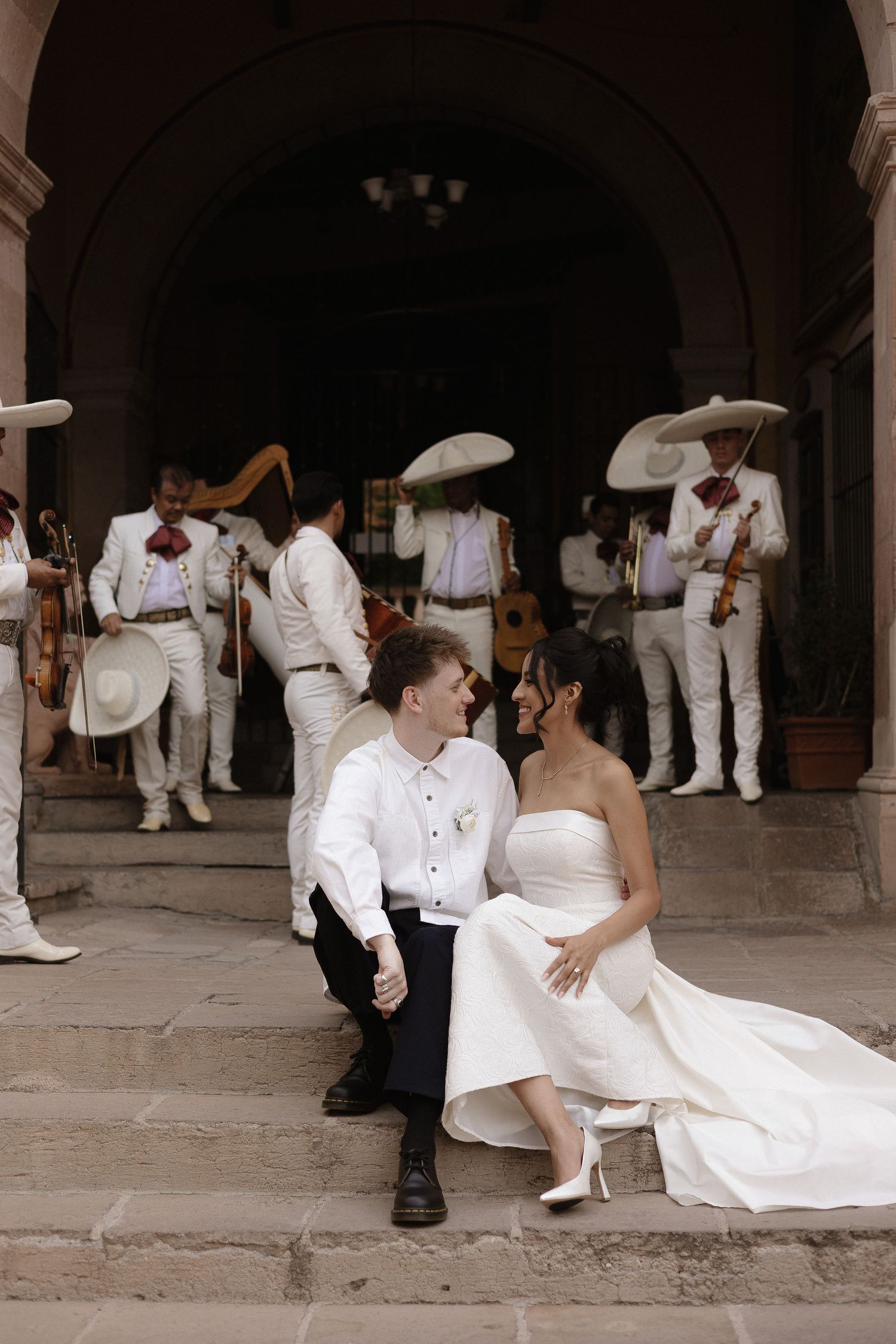 A wedding couple sits together on stone steps surrounded by guests dressed in white at an outdoor venue.