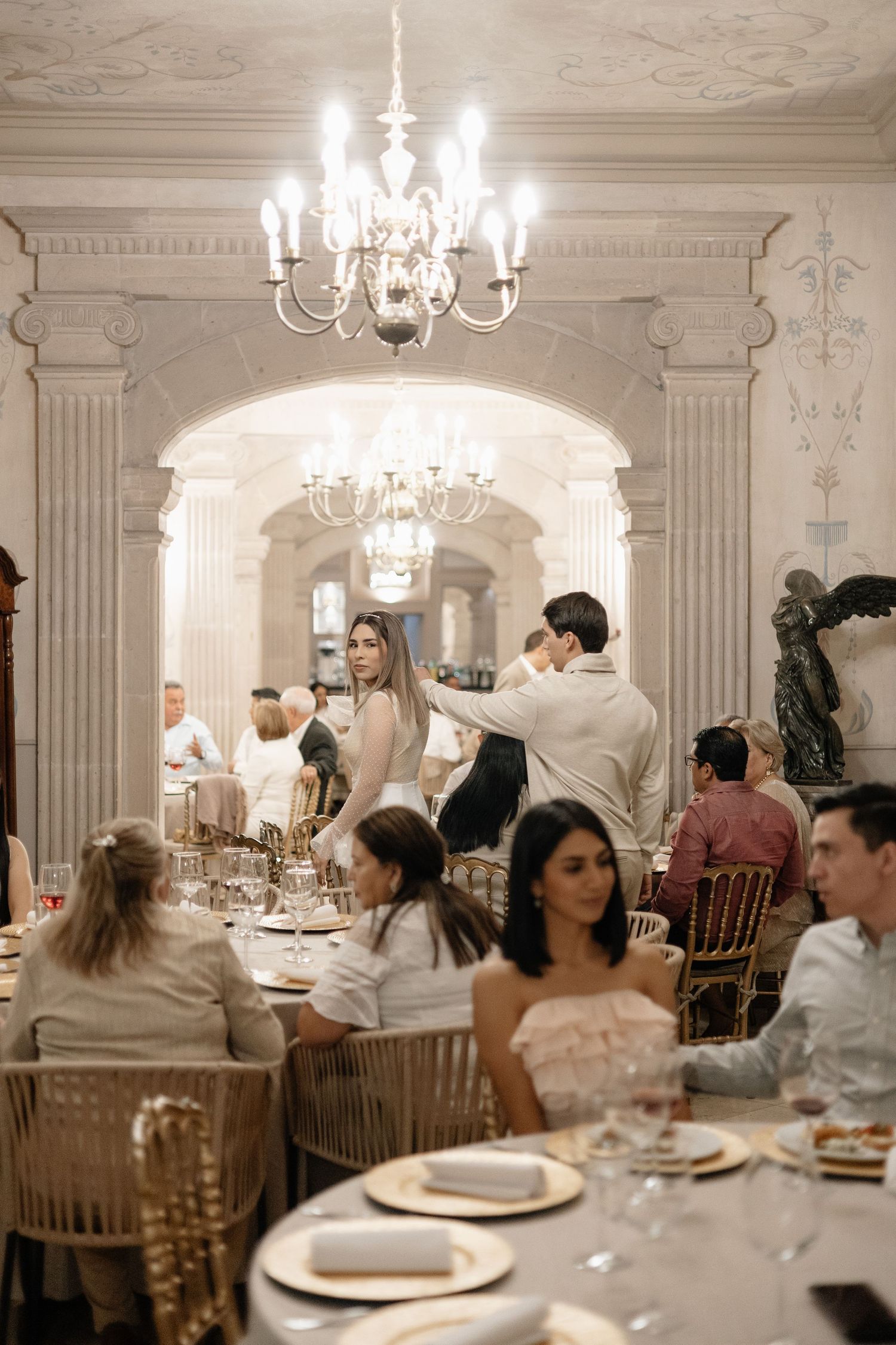 Elegant wedding reception in ornate white room with crystal chandeliers and guests seated at round tables.