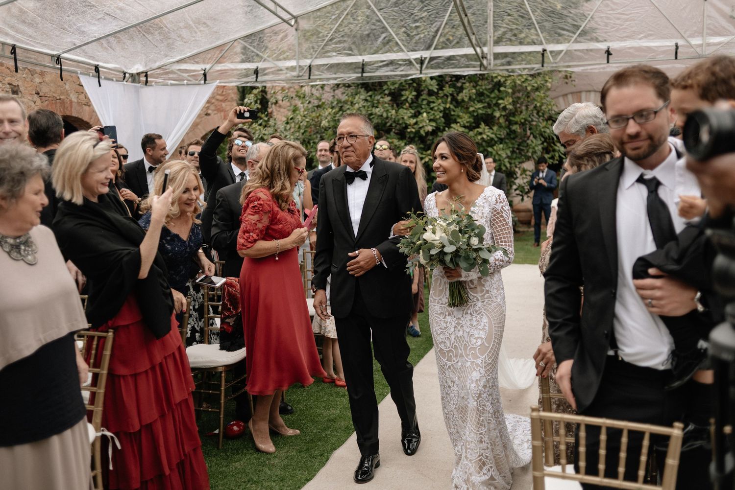 Wedding guests celebrate as newlyweds walk down the aisle in a elegant clear-tent outdoor reception.