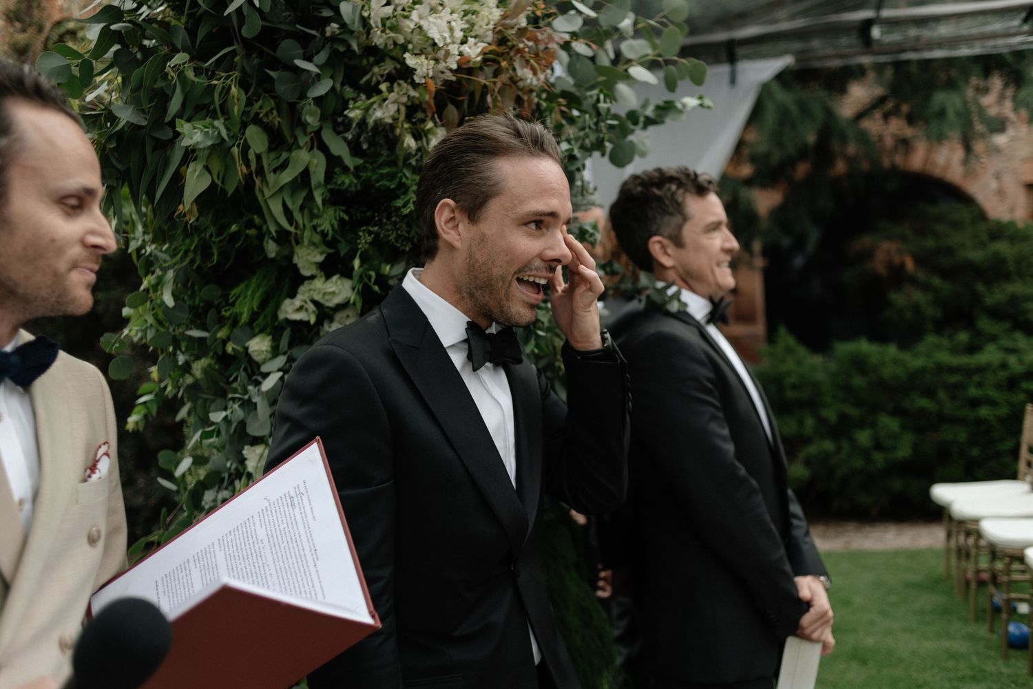 Three groomsmen in black tuxedos and bow ties stand together at an outdoor wedding ceremony venue.