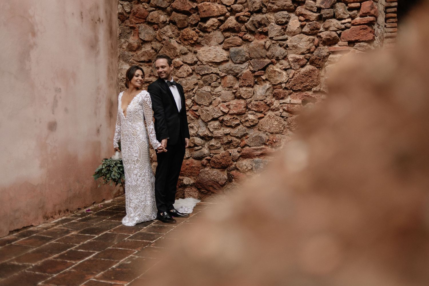 A bride and groom standing against a rustic stone wall in formal wedding attire.