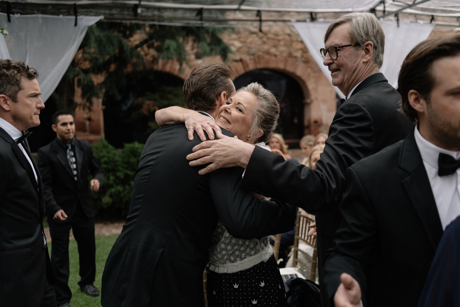 A couple embraces during a formal outdoor wedding ceremony while guests in black suits stand nearby.