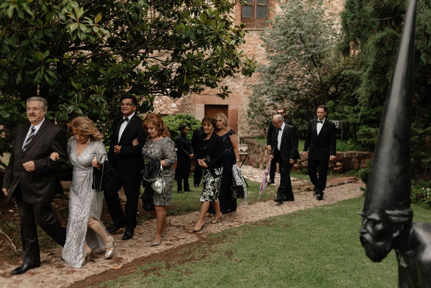 A wedding party walks along a garden path next to a brick building on a formal occasion.