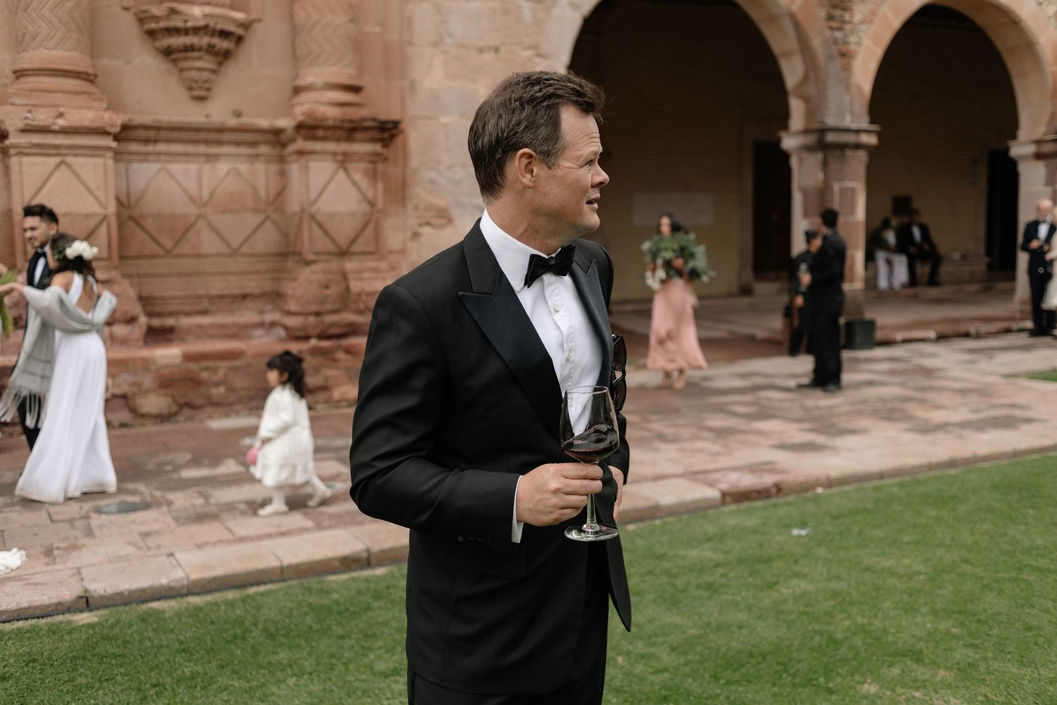 Groom in black tuxedo and bow tie holds drink glass while standing on lawn near arched stone building.
