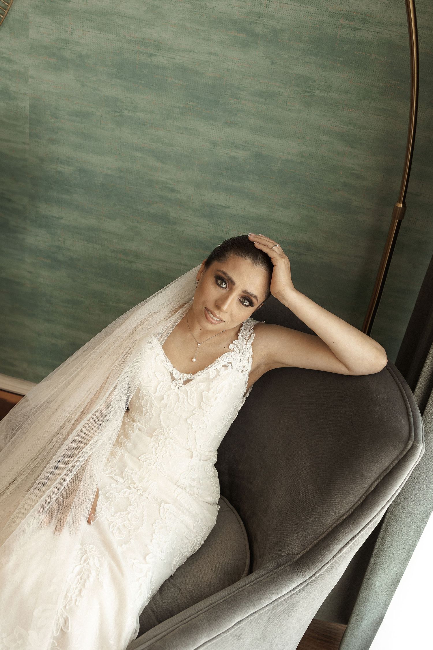 Bride wearing a fitted lace wedding dress and veil lounges on a dark couch against a green wall.