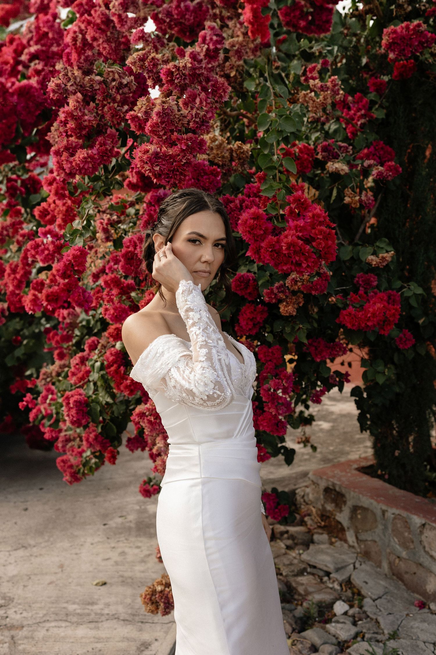 A figure in a white lace dress poses gracefully against a backdrop of vibrant red bougainvillea flowers in bloom.