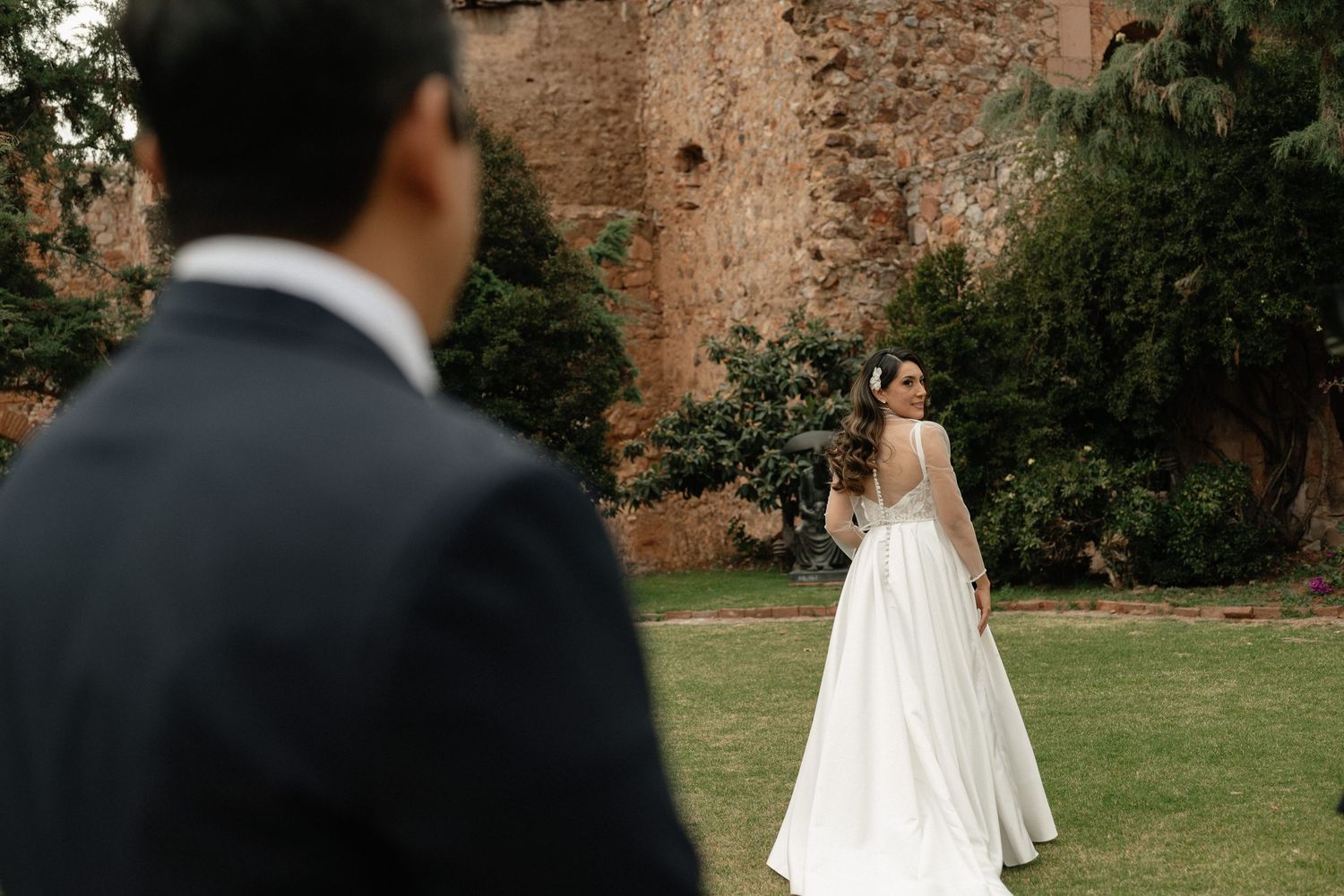 Newlyweds walk together on a lush green lawn beside historic brick walls during their outdoor wedding ceremony.