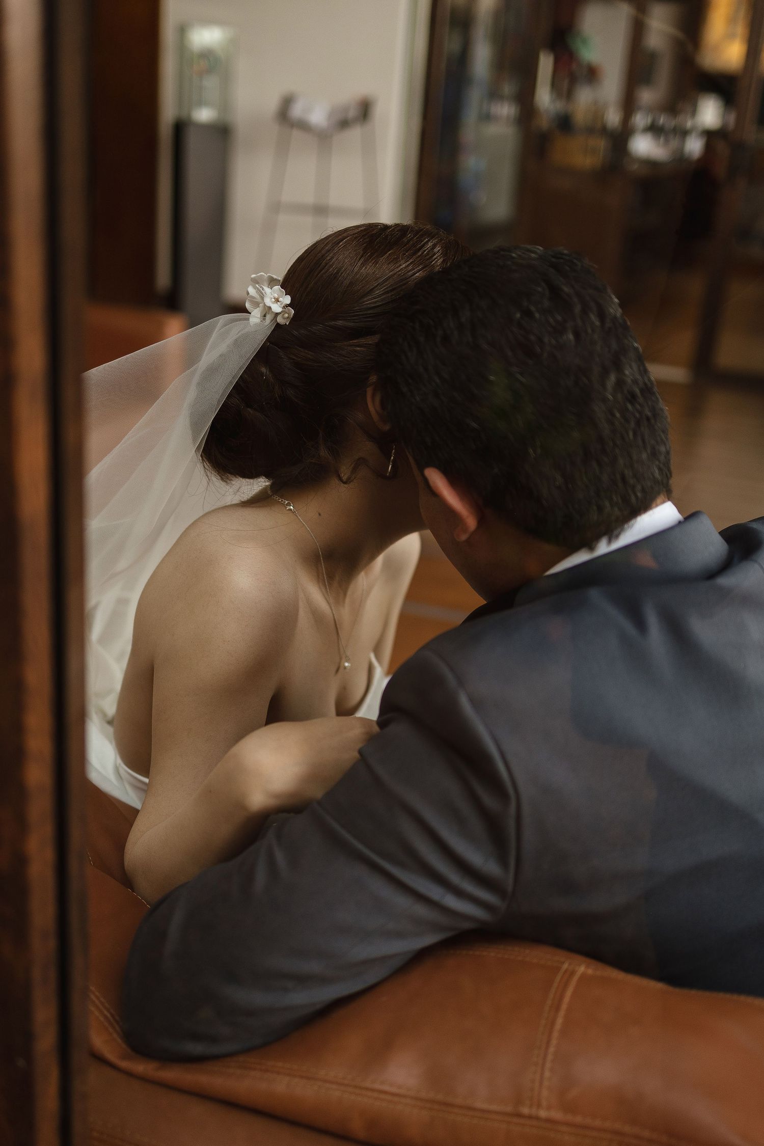 A tender moment between newlyweds as they sit together on a brown leather couch, the bride wearing a white veil.