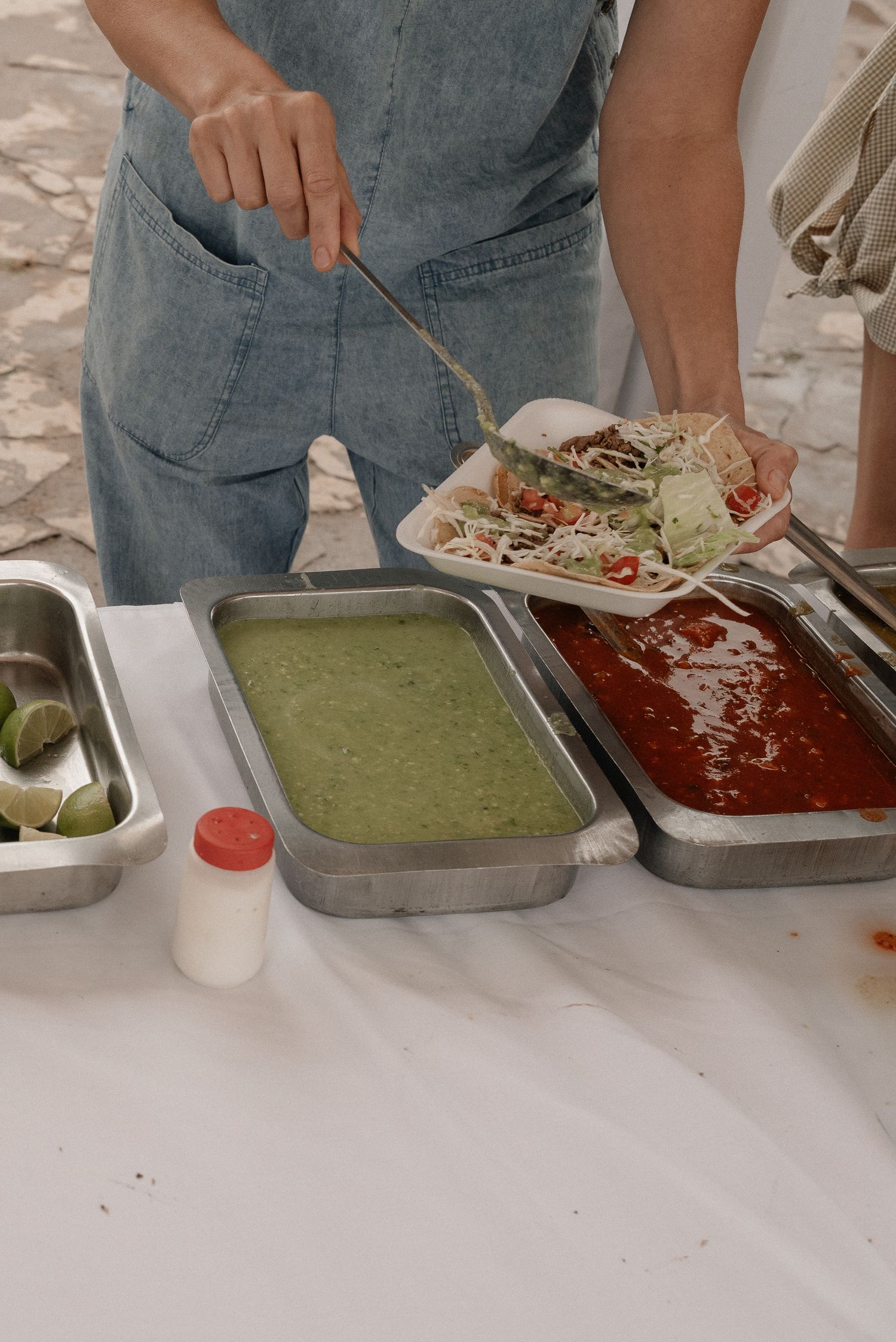 Person preparing Mexican food with red and green salsas, lettuce and limes in metal serving trays.