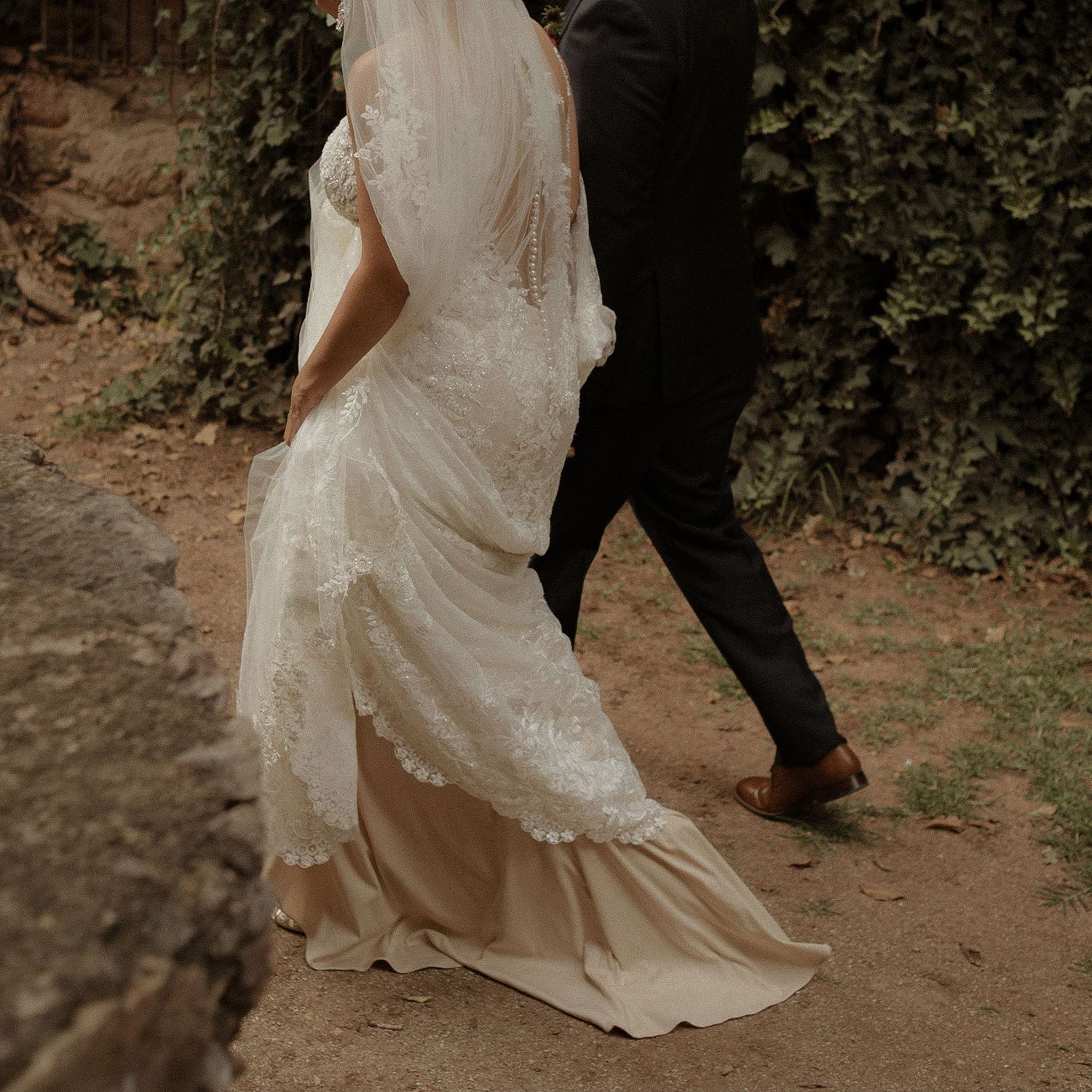Close-up of a bride's lace wedding dress trailing behind as couple walks on a path.