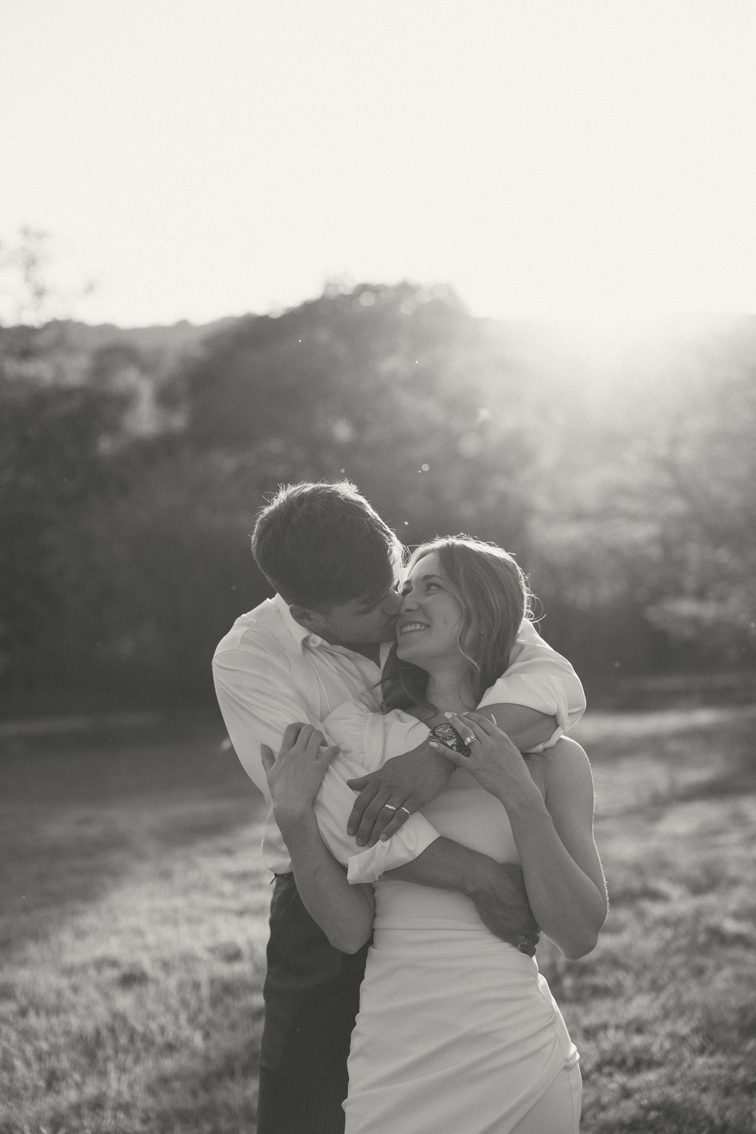 A romantic black and white photo of a couple embracing in a sunlit field at golden hour.