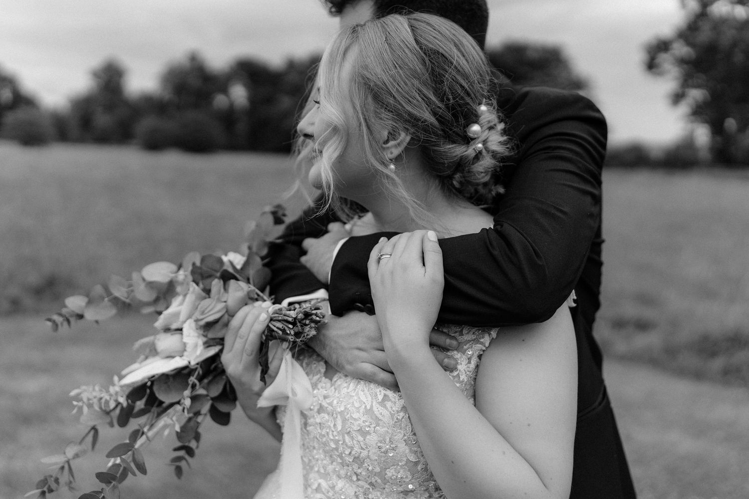 Black and white photo of a couple embracing in a field during their wedding, with the bride holding a floral bouquet.