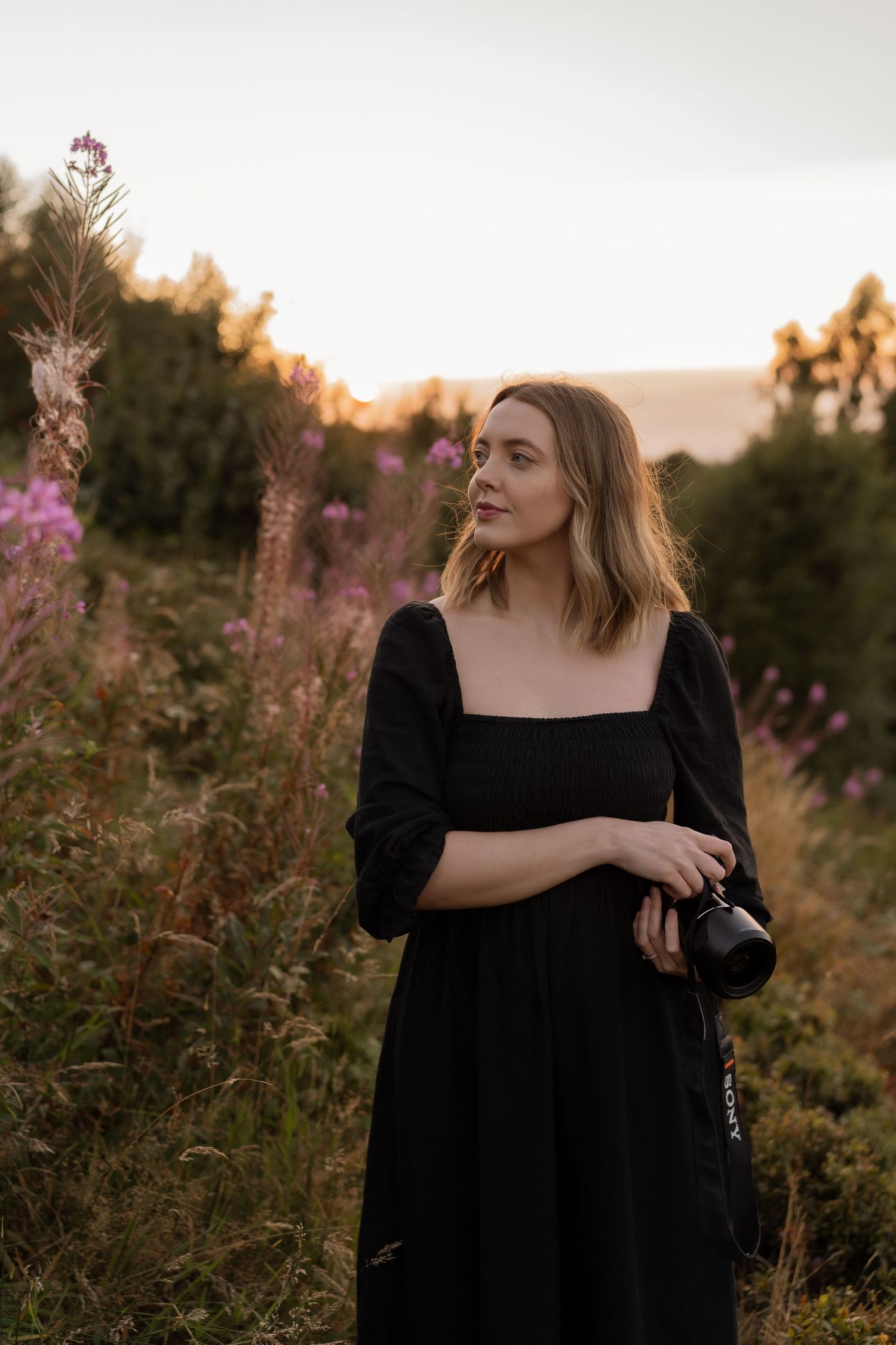 A person in a black dress poses in a wildflower field at sunset with purple blooms and golden light.