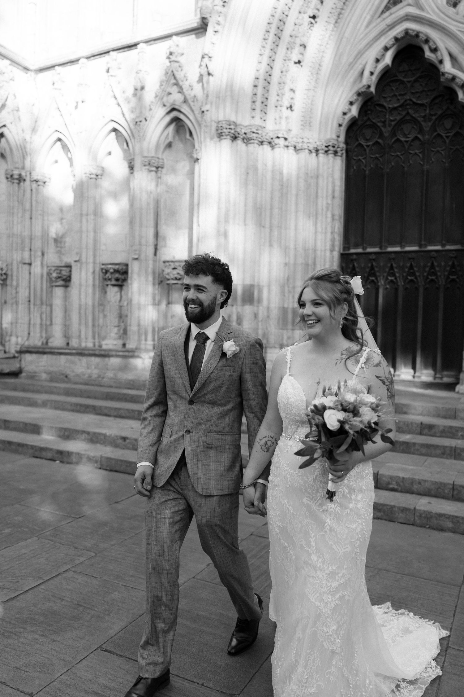 A happy couple walks together outside a Gothic cathedral after their wedding ceremony, captured in black and white.