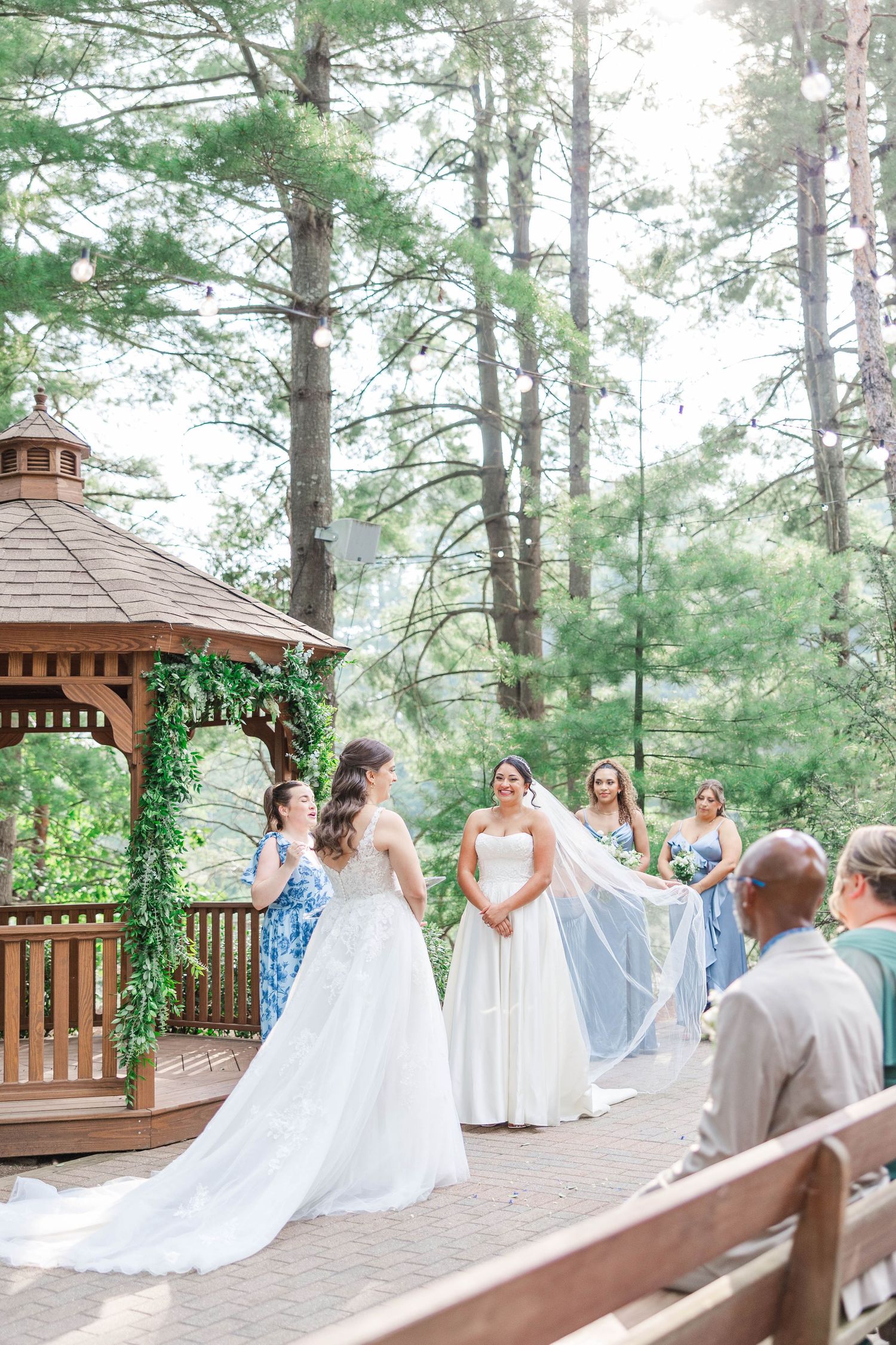 Wedding party in white dresses gathered on a wooden deck in a pine forest setting.