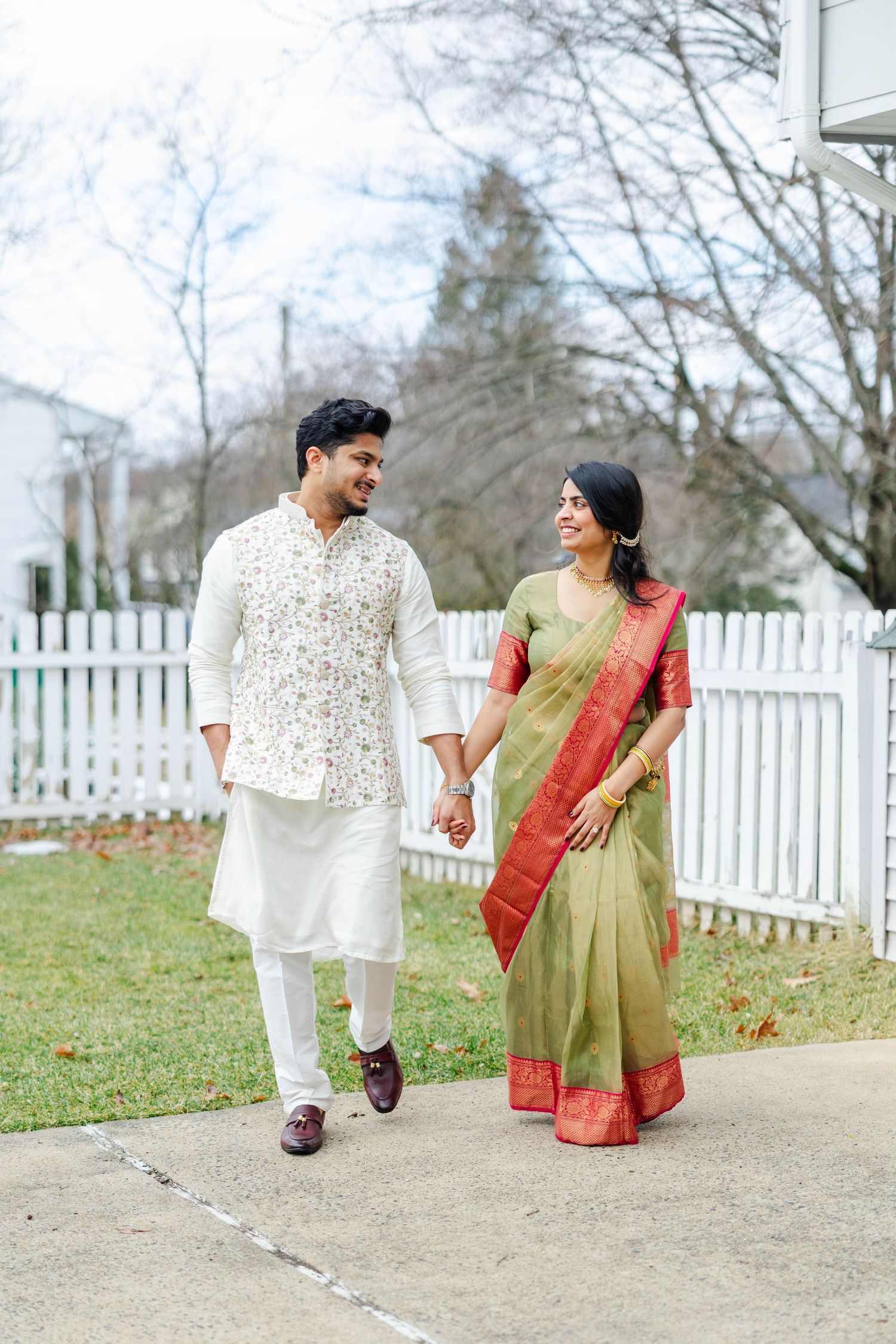 A couple in traditional Indian attire walking and laughing together along a white picket fence path in a garden setting.