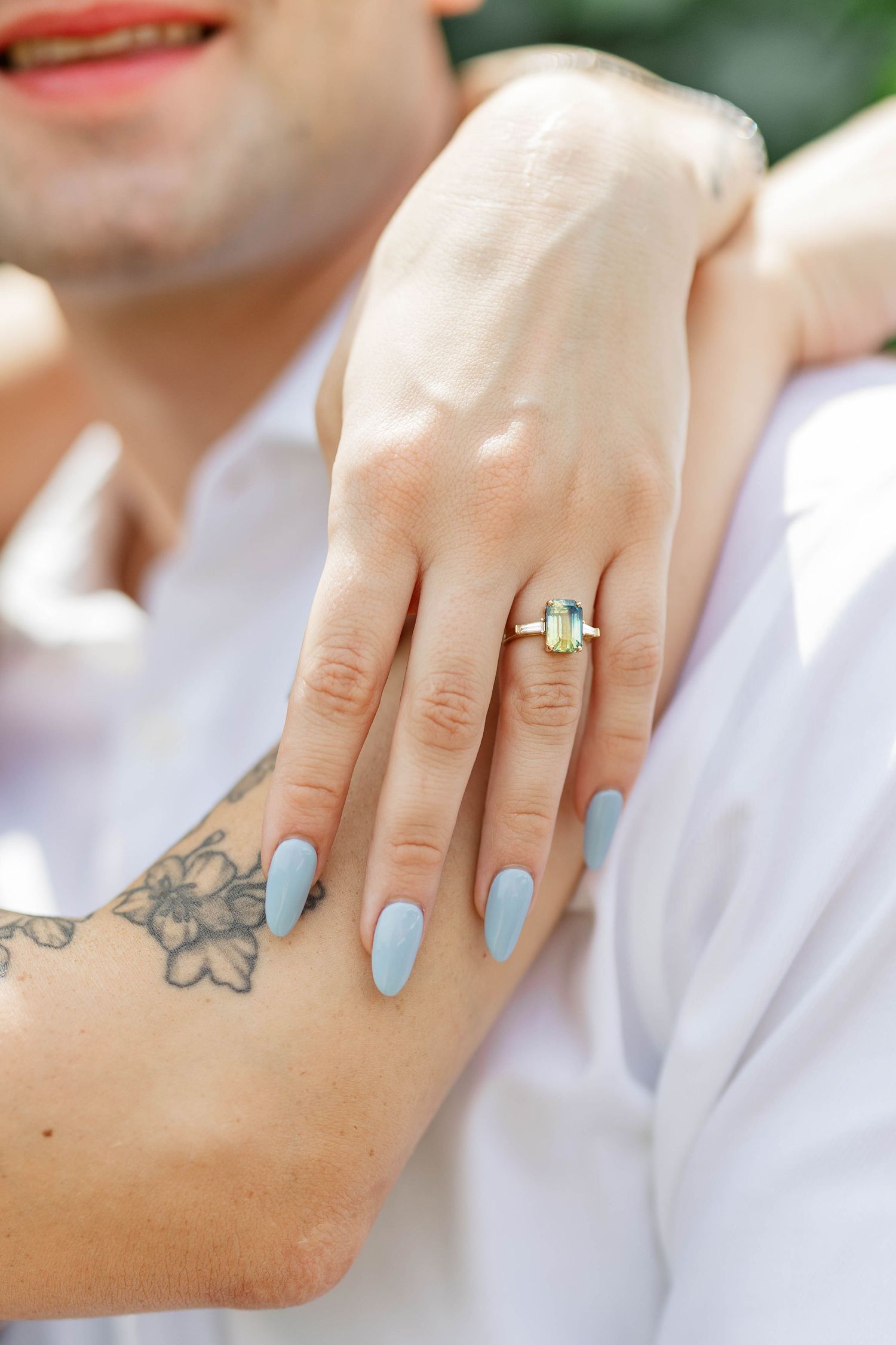 Woman with blue nails and yellow gemstone engagement ring resting her hand on a man's tattooed arm.