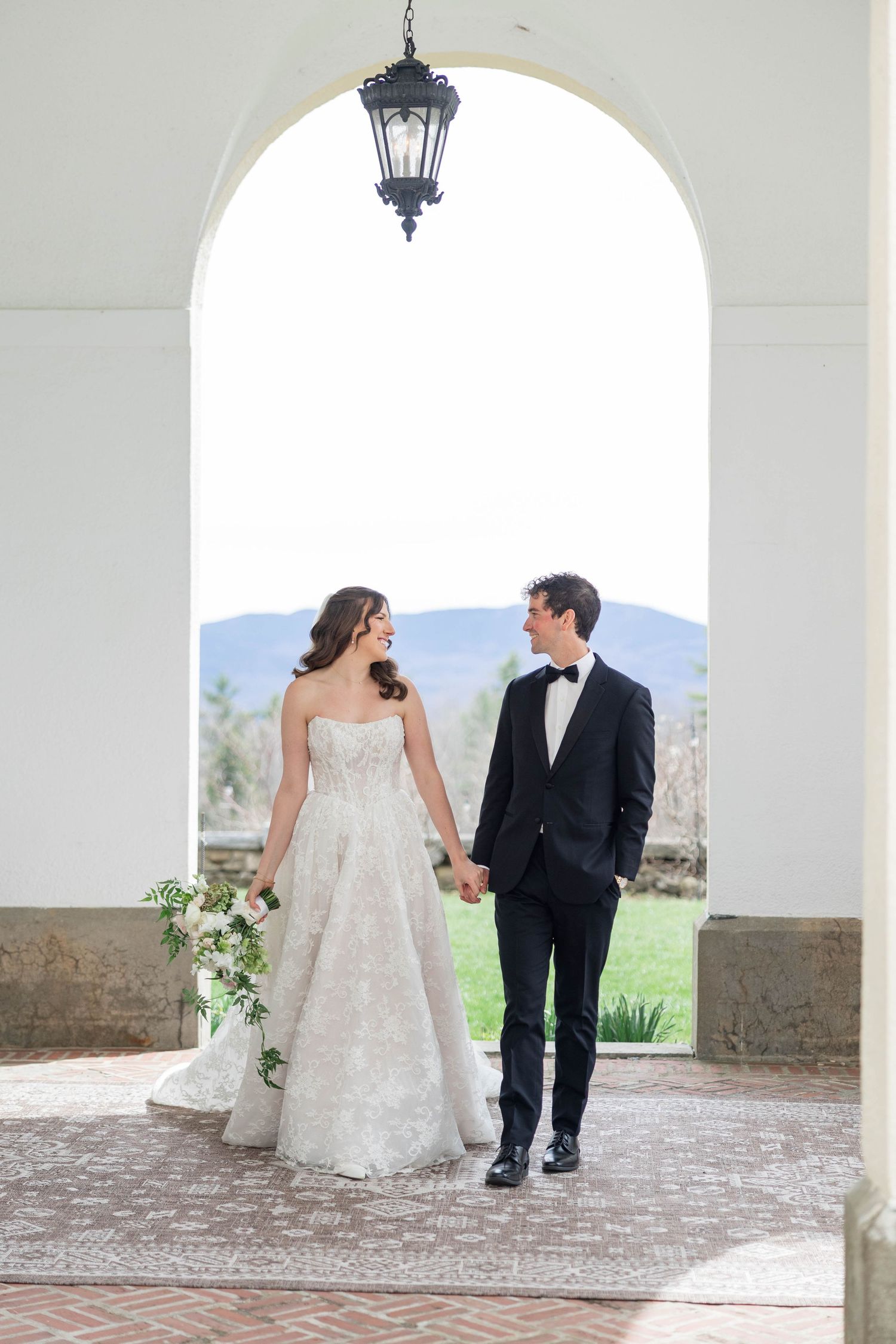 Bride and groom hold hands walking under elegant arch with mountain views on their wedding day.