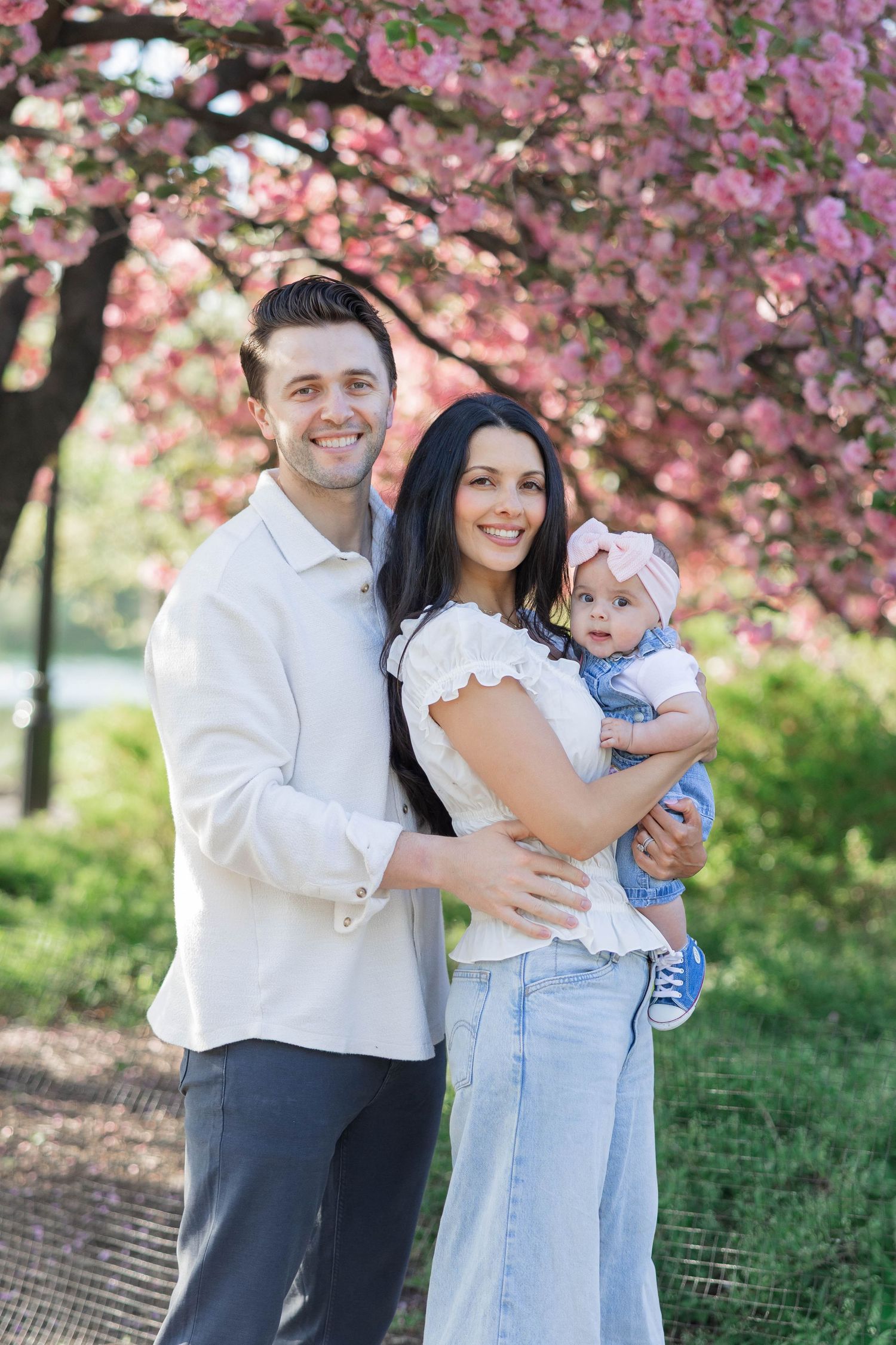 A smiling couple holds their baby girl under a blooming pink cherry blossom tree in a sunny park.