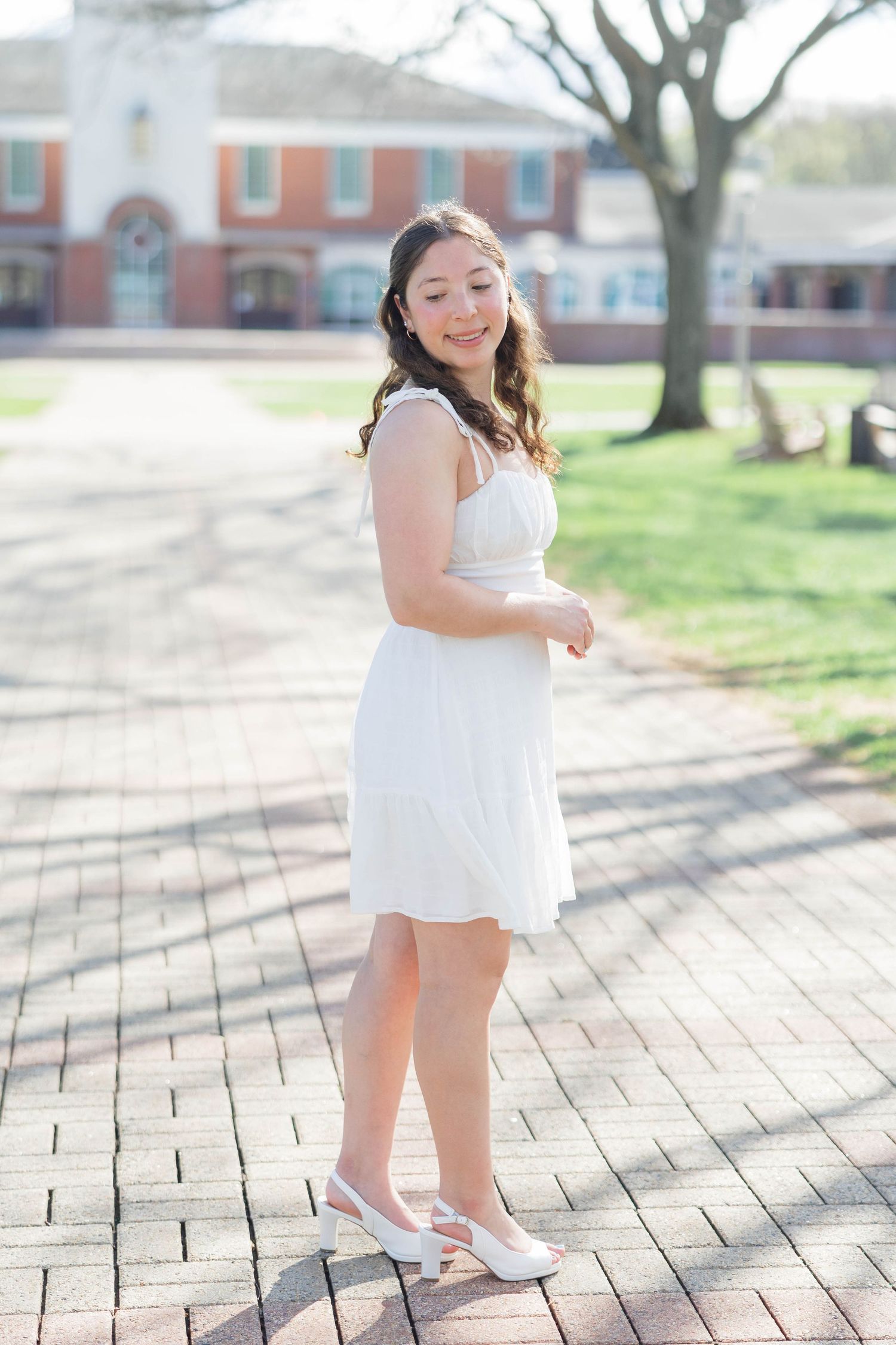 Young woman in white dress and white flats poses on brick pathway outside a red brick building on a sunny day.