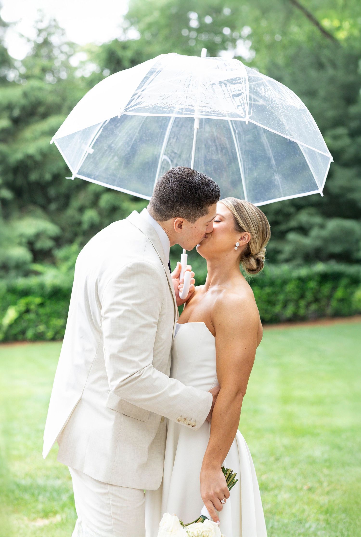 A romantic couple shares a kiss under a clear umbrella on a lush green lawn during their wedding celebration.