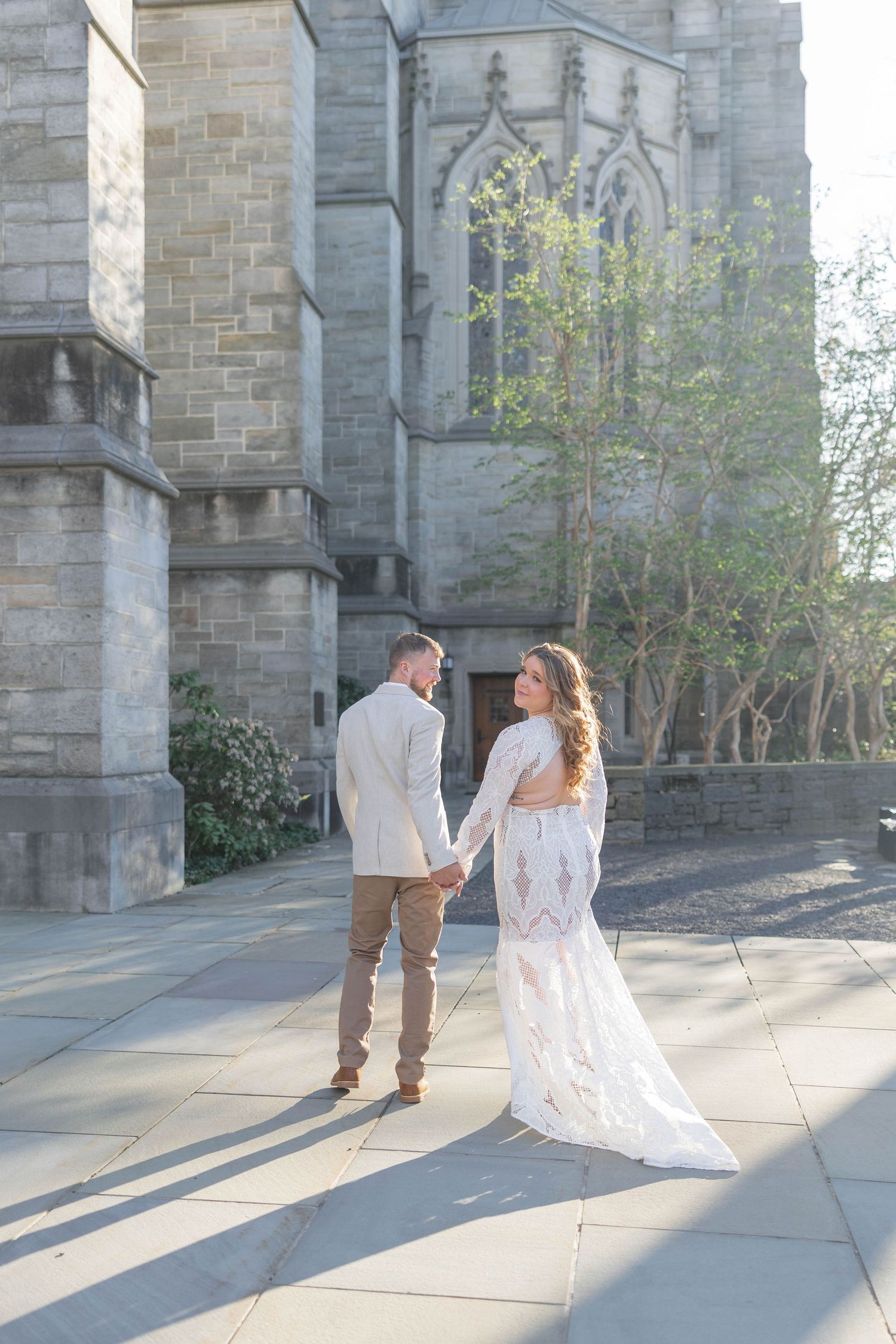 Bride in lace gown and groom in tan suit hold hands walking near a stone cathedral on a sunny day.