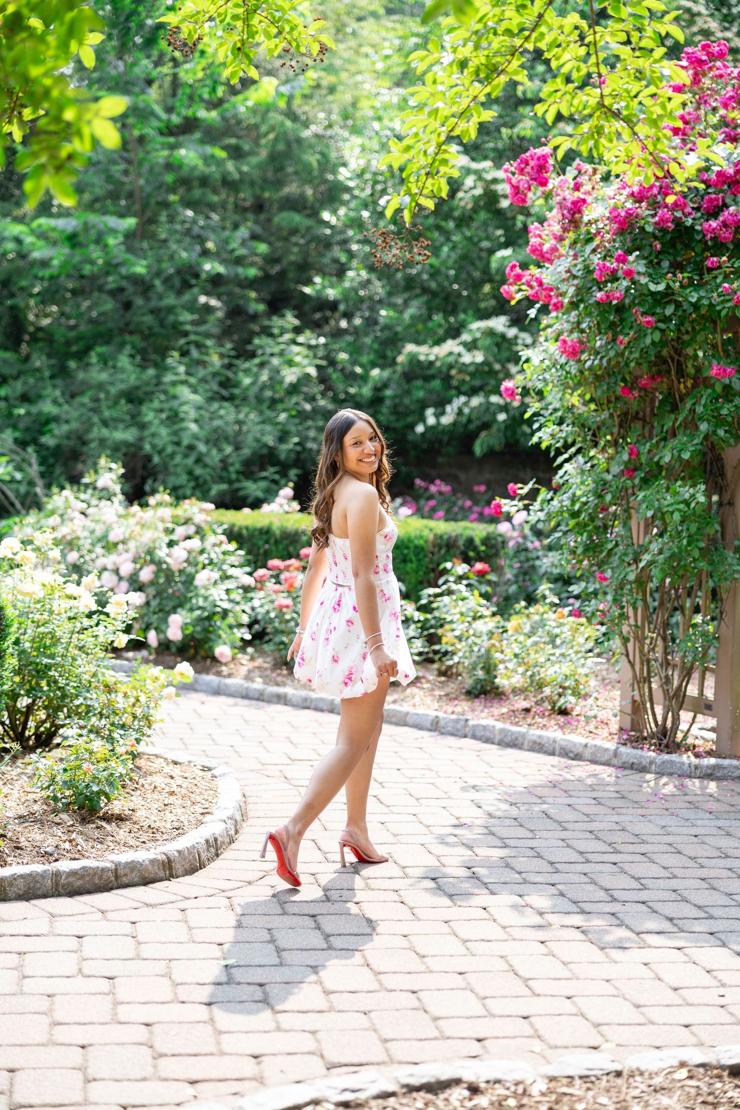 Woman in floral dress and red heels walking on brick path in lush garden with pink blooming flowers.
