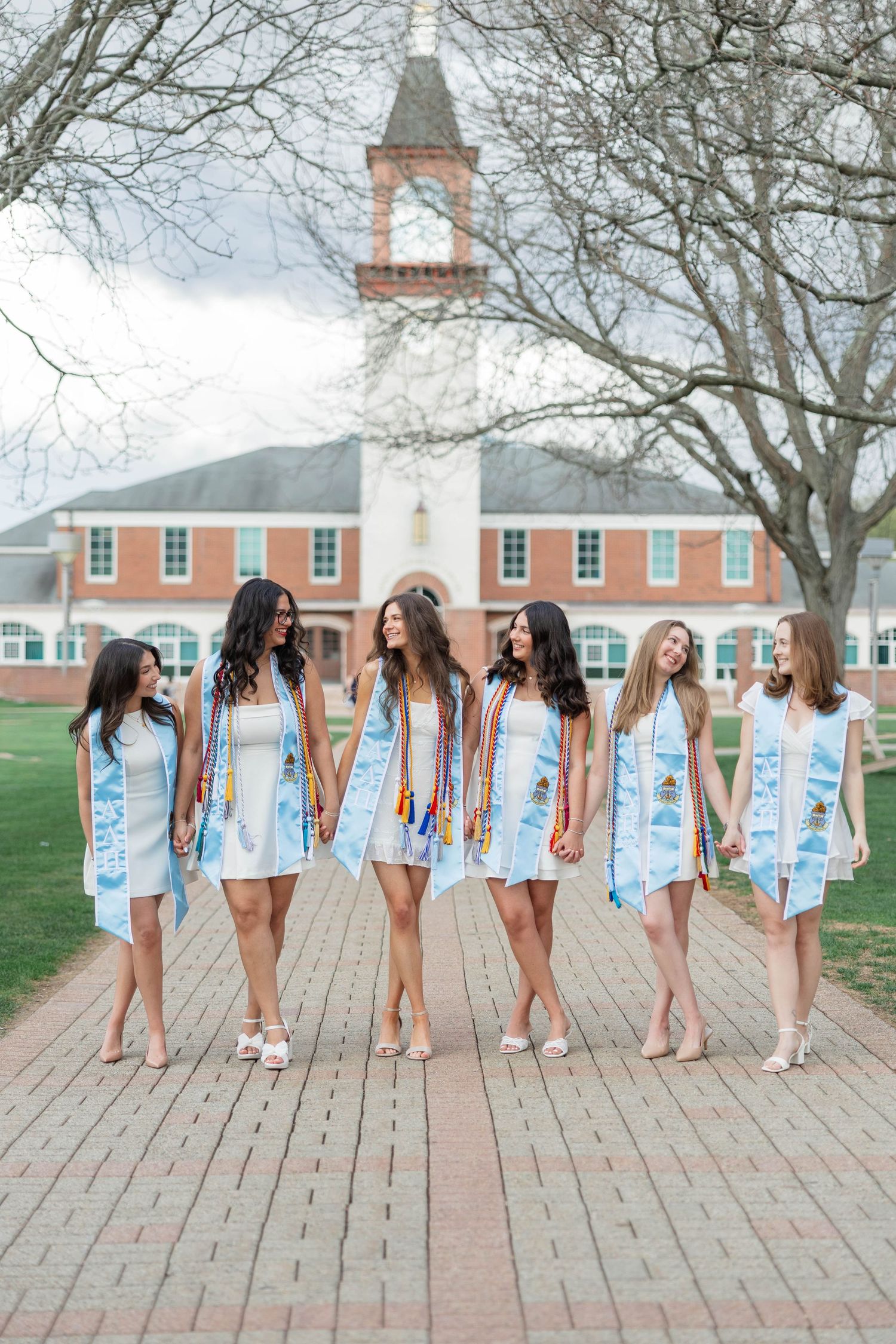 Six young women in white dresses and graduation stoles walk together on a brick path outside a brick university building.
