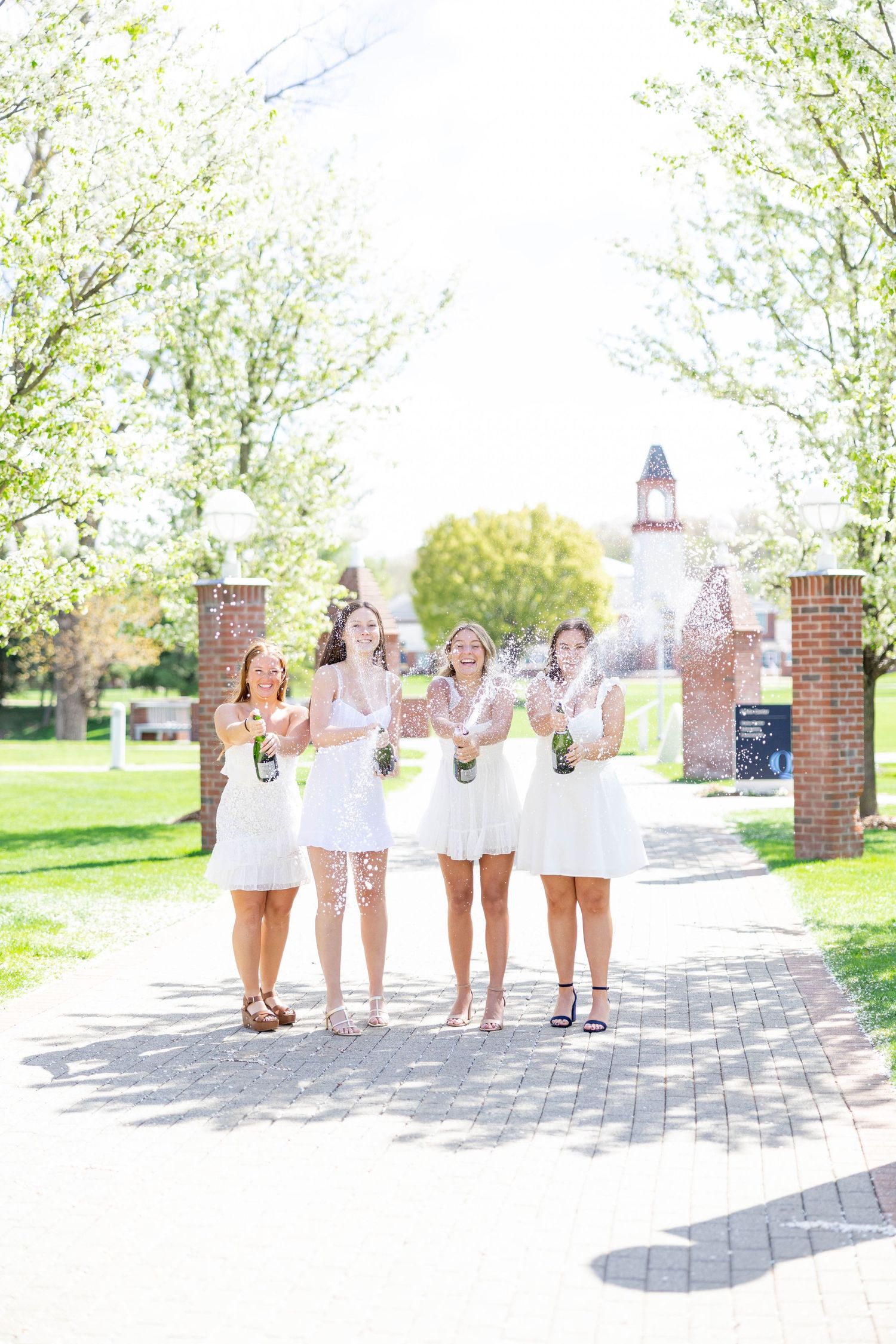 Four women in white dresses celebrate outdoors, popping champagne bottles on a sunny tree-lined brick pathway.
